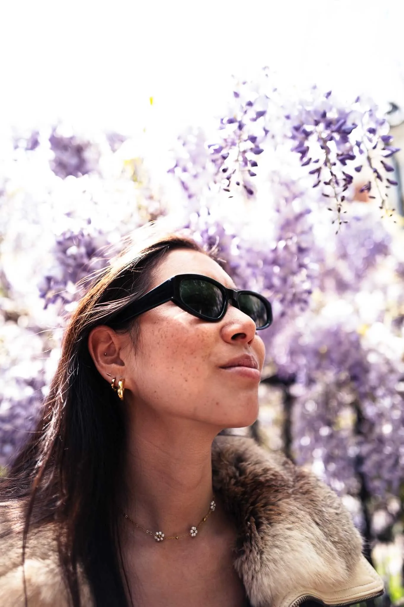 A woman wearing dark sunglasses and gold jewelry looks upward with her face tilted toward the sky, surrounded by blooming purple flowers that create a dreamy, overexposed background