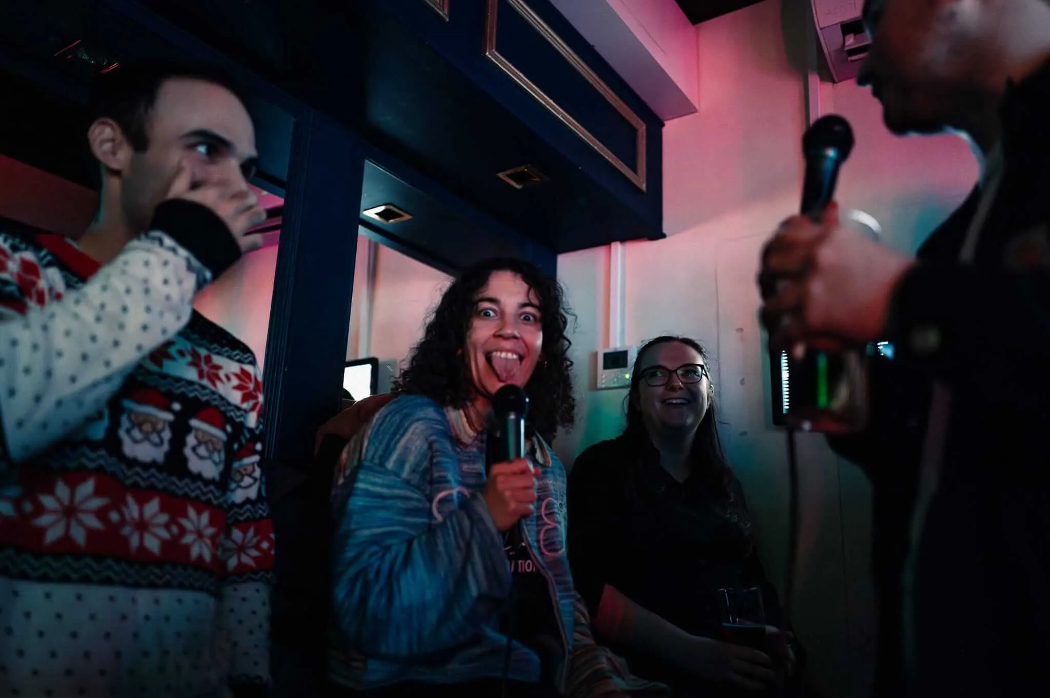 A group of friends gathered in a dimly lit karaoke room, with one woman in the center holding a microphone and making an exaggerated facial expression with her tongue out, while others smile and laugh around her. The scene is bathed in pink and blue neon lighting typical of karaoke venues.