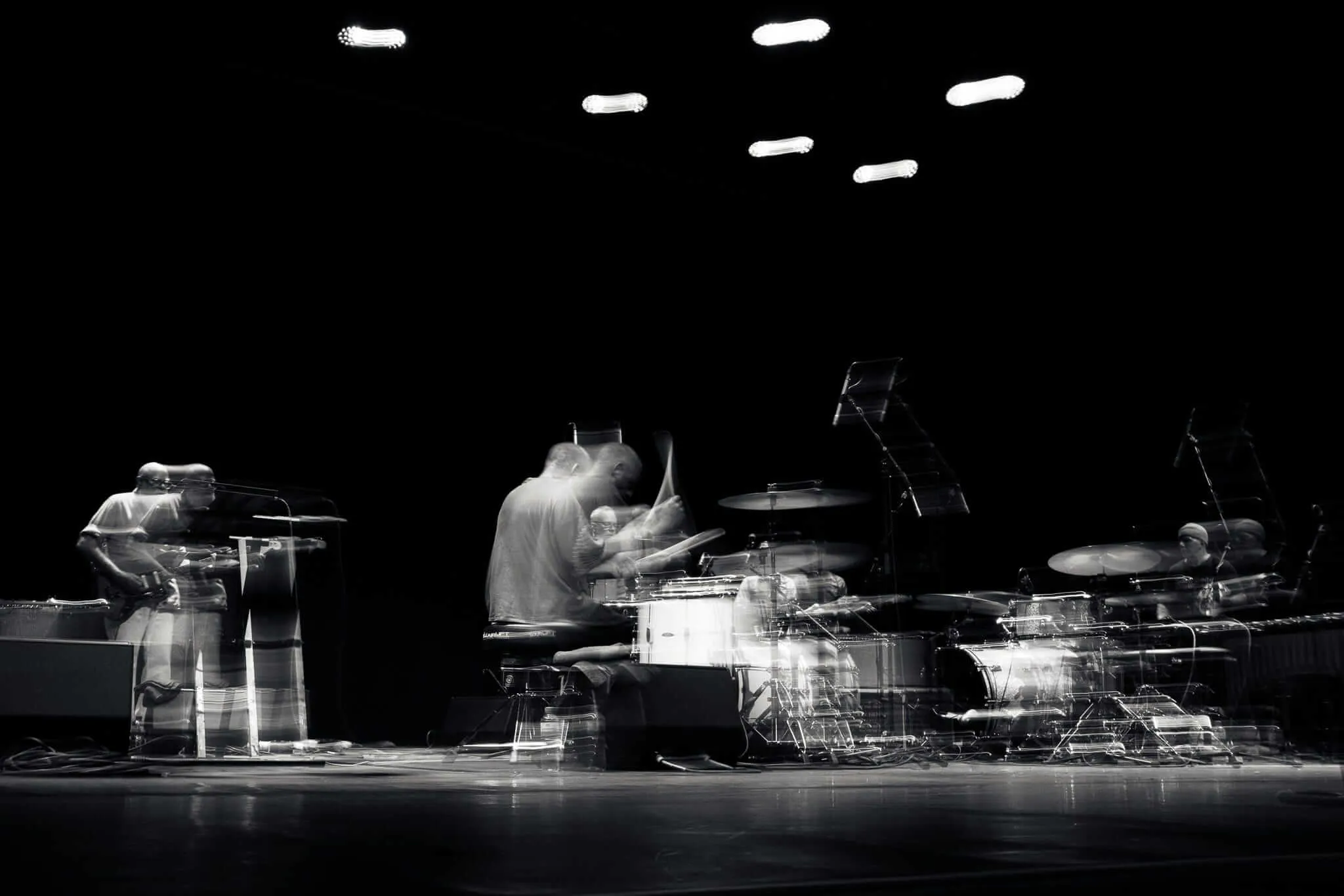 Black and white photograph of musicians performing on stage, with a drummer in the center, captured with motion blur that creates ghostly, ethereal effects against the dark background with stage lights visible above