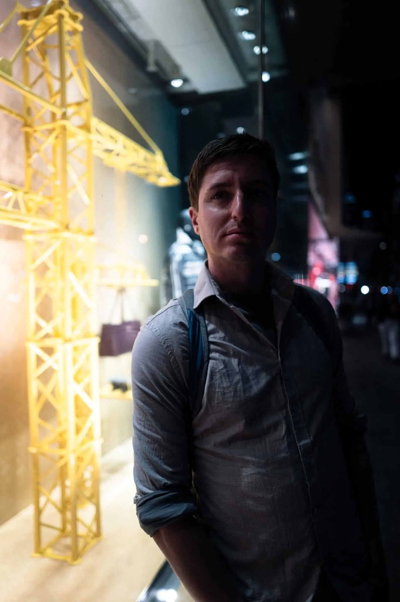 A young man in a grey button-up shirt stands before a glowing yellow crane model display in a dimly lit museum or gallery window, his face partially lit by the warm exhibition light as he gazes upward with a contemplative expression.
