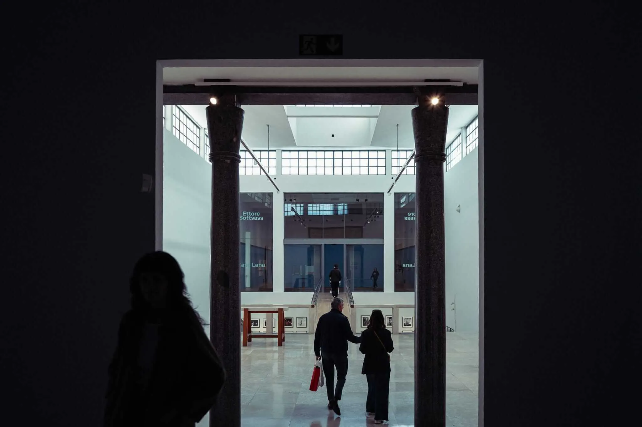 A silhouetted figure in the foreground watches as two visitors walk through a grand doorway flanked by granite columns into a brightly lit museum atrium displaying an Ettore Sottsass exhibition, with stairs and glass walls visible in the background.
