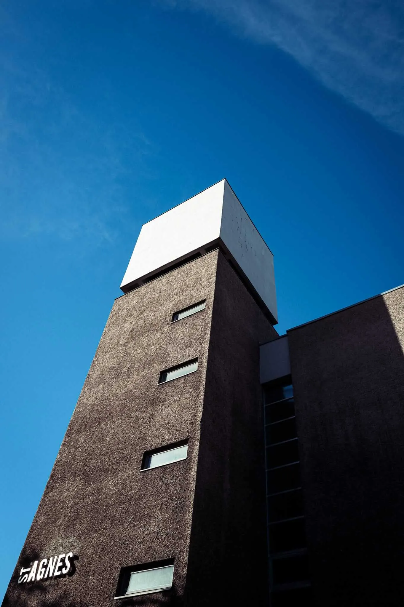 A dramatic low-angle view of a modern building with a dark textured facade featuring horizontal strip windows on multiple floors, topped by a distinctive white geometric crown structure against a brilliant blue sky. The word 'ST AGNES' is visible on the building's lower portion.