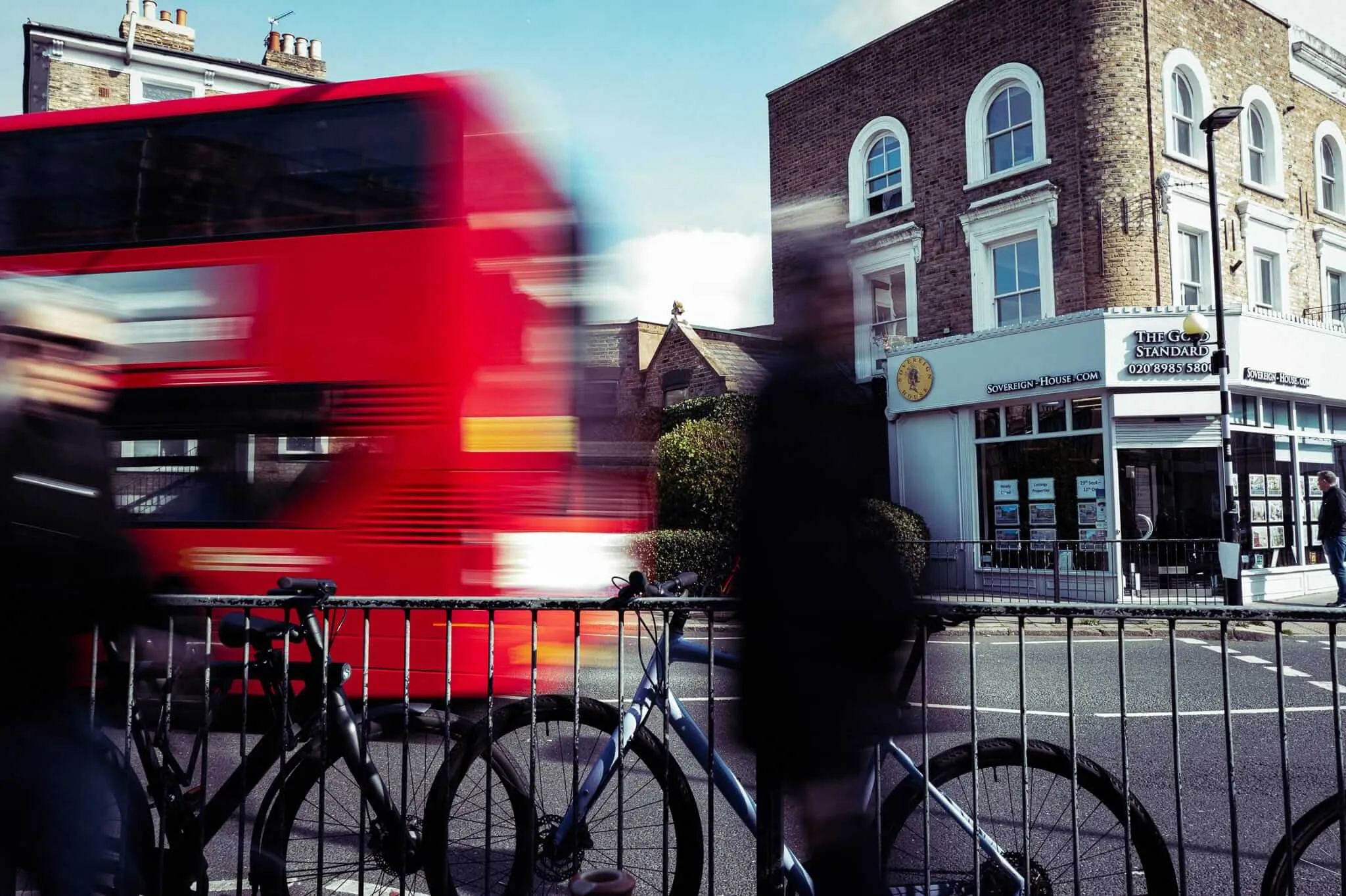 A blurred red double-decker bus speeds past Victorian brick buildings while bicycles are parked behind metal railings in the foreground. The motion blur creates streaks of red and white against the static urban architecture.