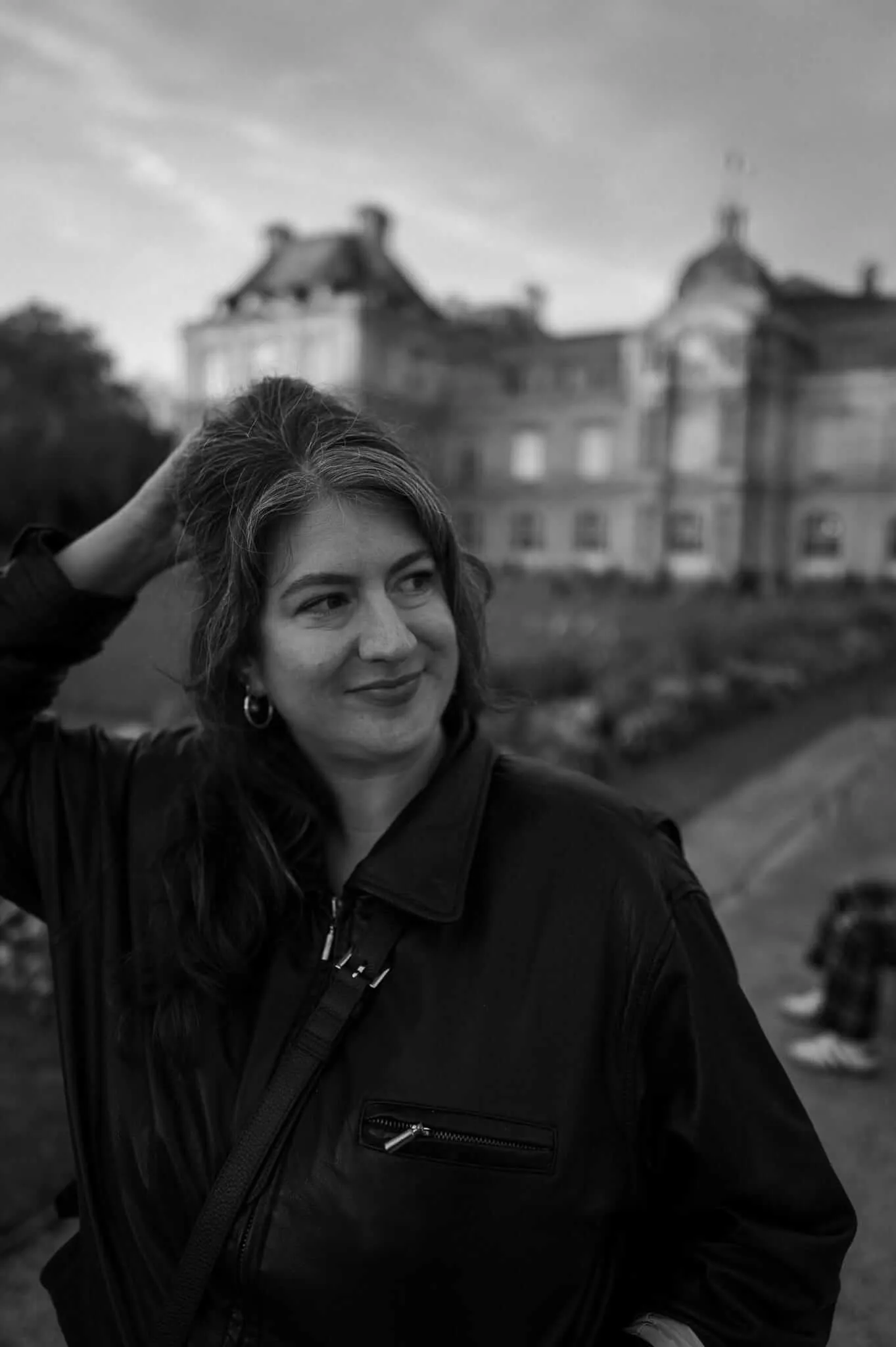 A black and white portrait of a smiling woman with her hand running through her hair, wearing a dark jacket with a cross-body bag strap visible. Behind her is a blurred classical European building with ornate architecture and a cloudy sky.
