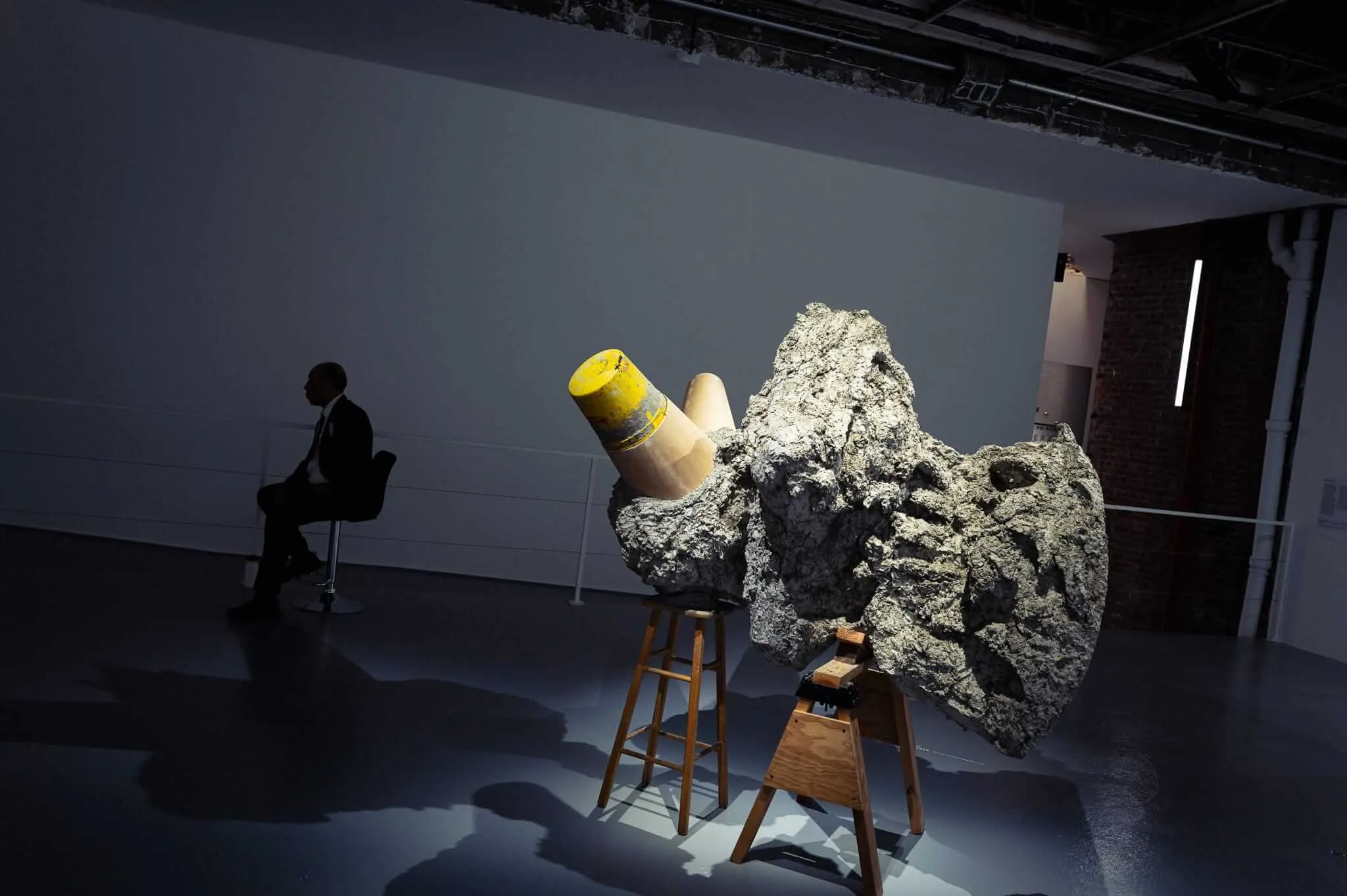 A contemporary art gallery scene showing a silhouetted figure in a suit sitting on a chair in the background, while in the foreground a large rocky sculptural piece mounted on wooden supports dominates the space, with yellow and beige cylindrical forms emerging from or embedded within the rough-textured stone mass