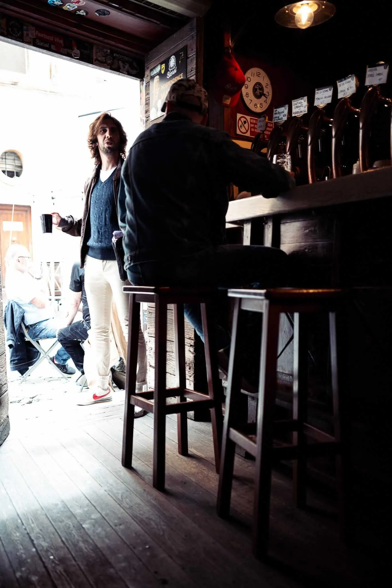 Interior of a dimly lit traditional pub with wooden bar stools and beer taps. A man in a blue sweater stands at the bar while another person sits on a stool facing away from camera. Warm lighting creates atmospheric shadows, with a clock visible on the wood-paneled wall and bright daylight streaming through windows.