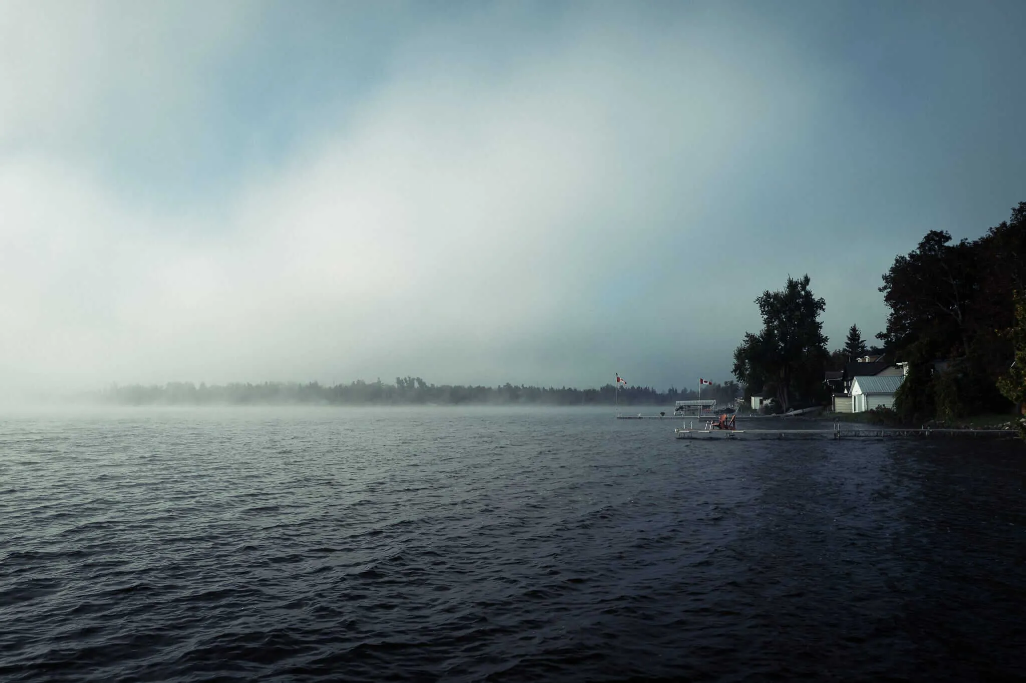 A misty morning over a dark, rippling lake with fog rolling across the far treeline. On the right shore, docks, cottages, and Canadian flags are partially visible among dense trees under an overcast sky.