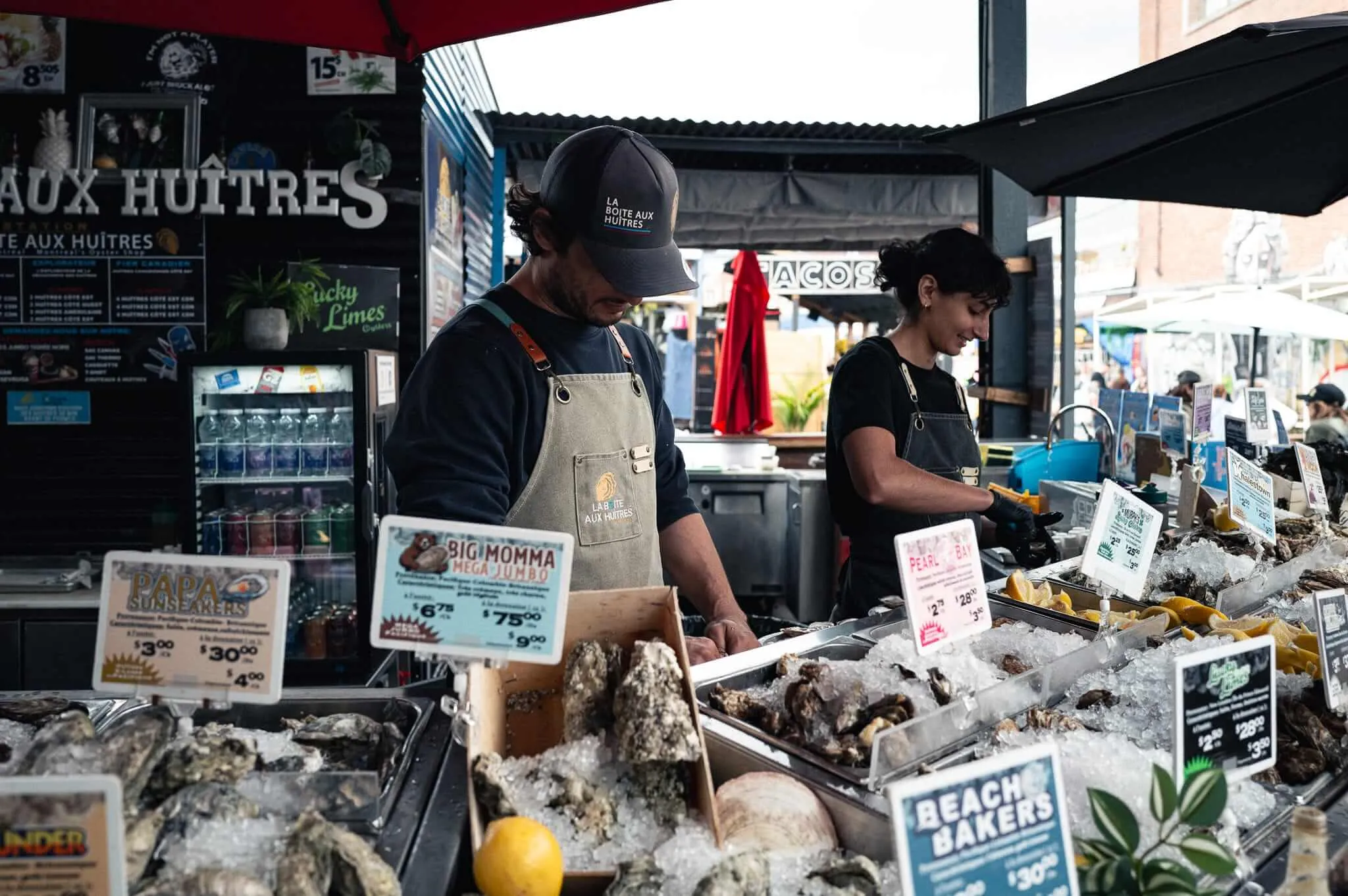 Two workers in aprons shuck oysters at an outdoor market stall called La Boîte aux Huîtres, with trays of fresh oysters on ice and price signs visible in the foreground.