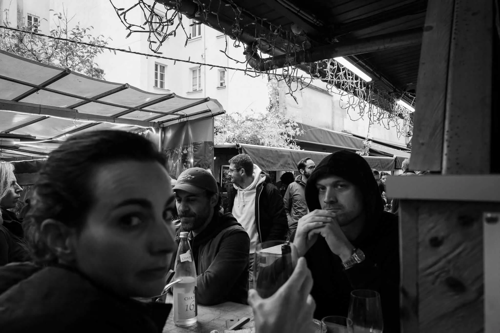 Black and white photograph of people gathered around tables under a covered outdoor area or market, with classical European architecture visible in the background. The scene captures intimate conversations and social interaction in what appears to be a café or restaurant setting.