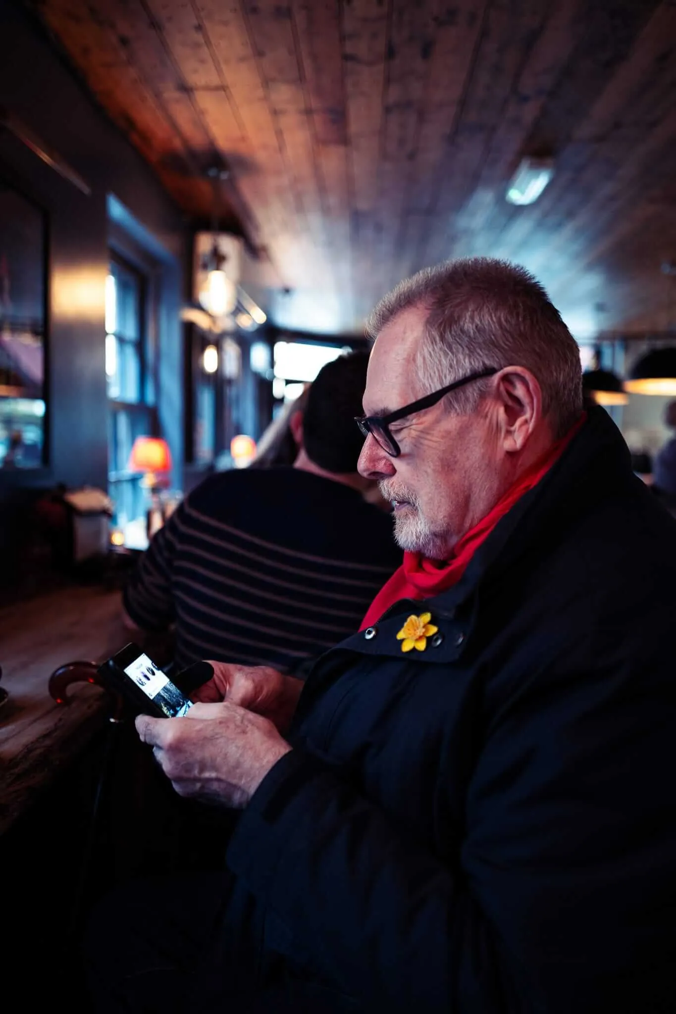 An elderly man with glasses and a red scarf sits in what appears to be a train or bus interior, looking down at his smartphone. The wooden-paneled ceiling and warm ambient lighting create an intimate atmosphere as he concentrates on his device during his journey.