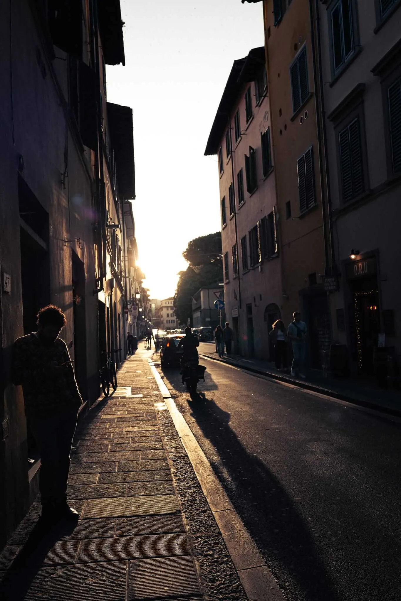 A narrow European street at golden hour with silhouetted figures walking along cobblestone sidewalks between tall buildings with shuttered windows, creating dramatic shadows and warm backlighting