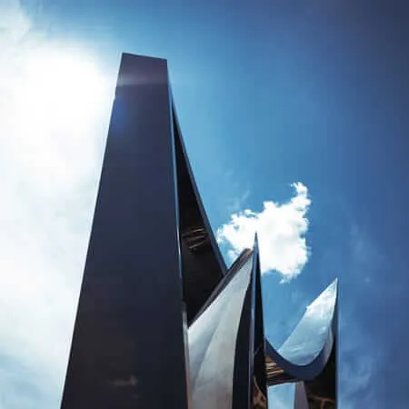 A low-angle view looking up at a towering abstract metal sculpture with sharp, blade-like forms and polished reflective surfaces, set against a vivid blue sky with scattered clouds and bright sunlight.