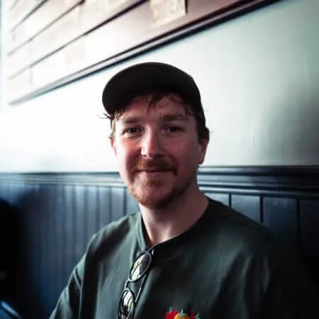 A bearded man wearing a black baseball cap and green Bodegas t-shirt stands in what appears to be a grocery store or market, with product shelves visible behind him. He has a slight smile and is looking directly at the camera.