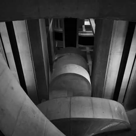 Black and white photograph taken from below showing a dramatic upward view of curved concrete steps or levels ascending in a spiral pattern, framed by vertical concrete pillars and glass panels, creating strong geometric shadows and architectural depth