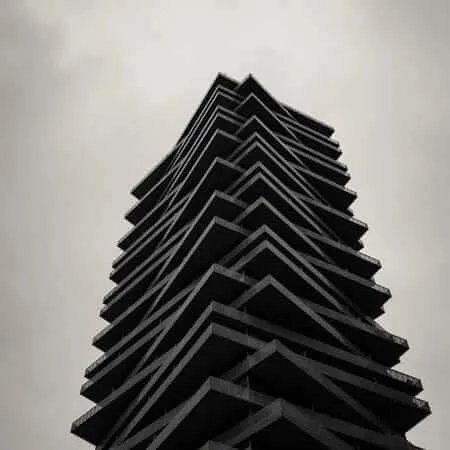 A dramatic low-angle black and white photograph of a modern high-rise tower with staggered, angular balconies that taper toward the top, resembling a ziggurat, shot against an overcast grey sky with a tree silhouette visible at the base.