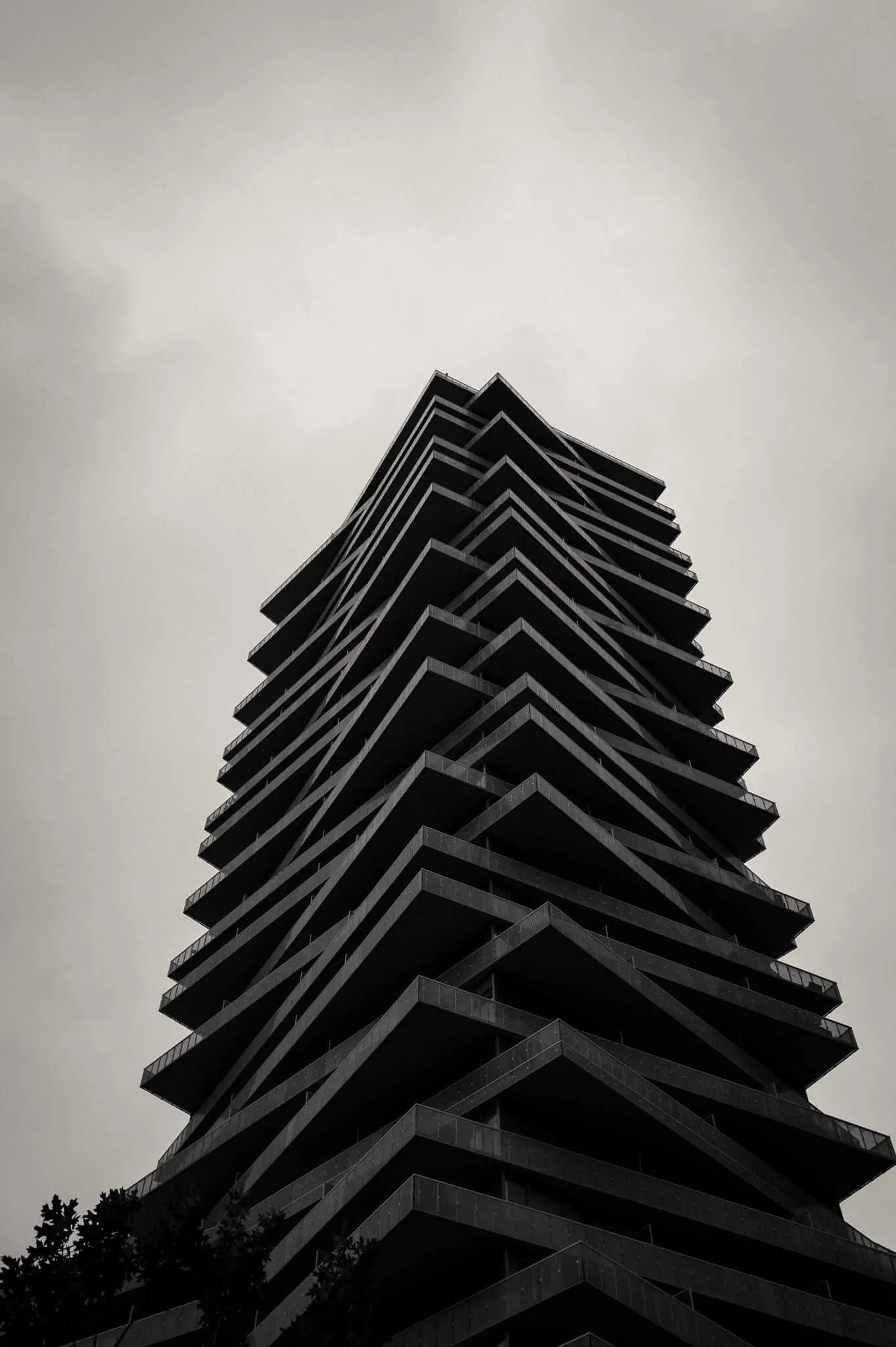A dramatic low-angle black and white photograph of a modern high-rise tower with staggered, angular balconies that taper toward the top, resembling a ziggurat, shot against an overcast grey sky with a tree silhouette visible at the base.