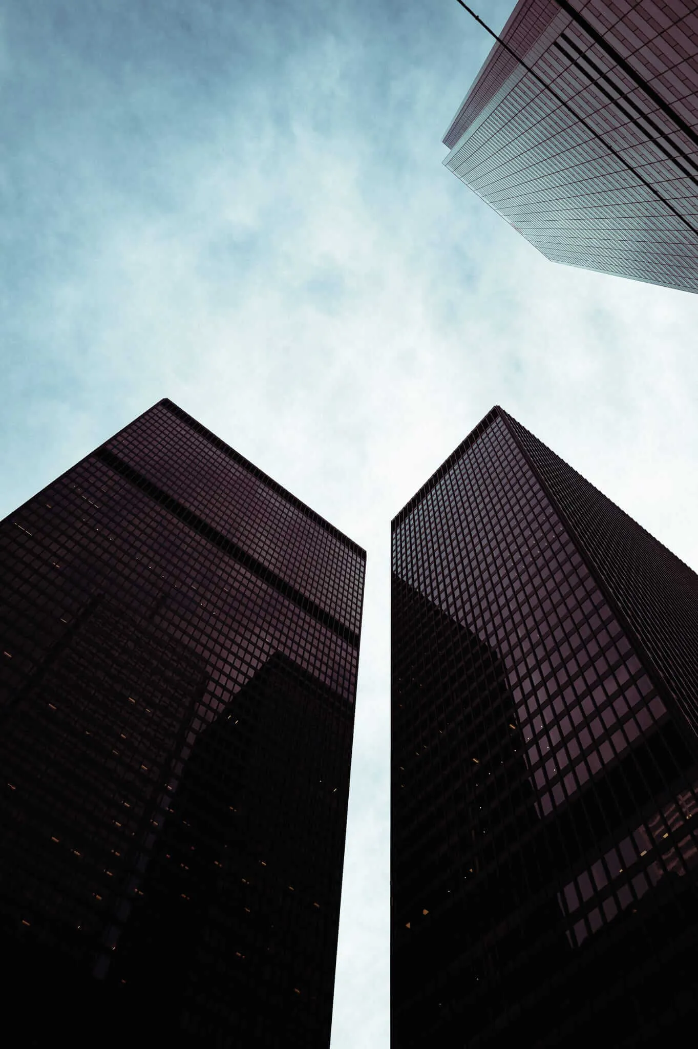 A dramatic low-angle upward perspective photograph of two dark glass and steel skyscrapers rising symmetrically against a pale blue cloudy sky, with a third glass tower visible in the upper right corner. The buildings are rendered in deep shadow, creating a stark silhouette effect, while a sliver of sky is visible between the two central towers.