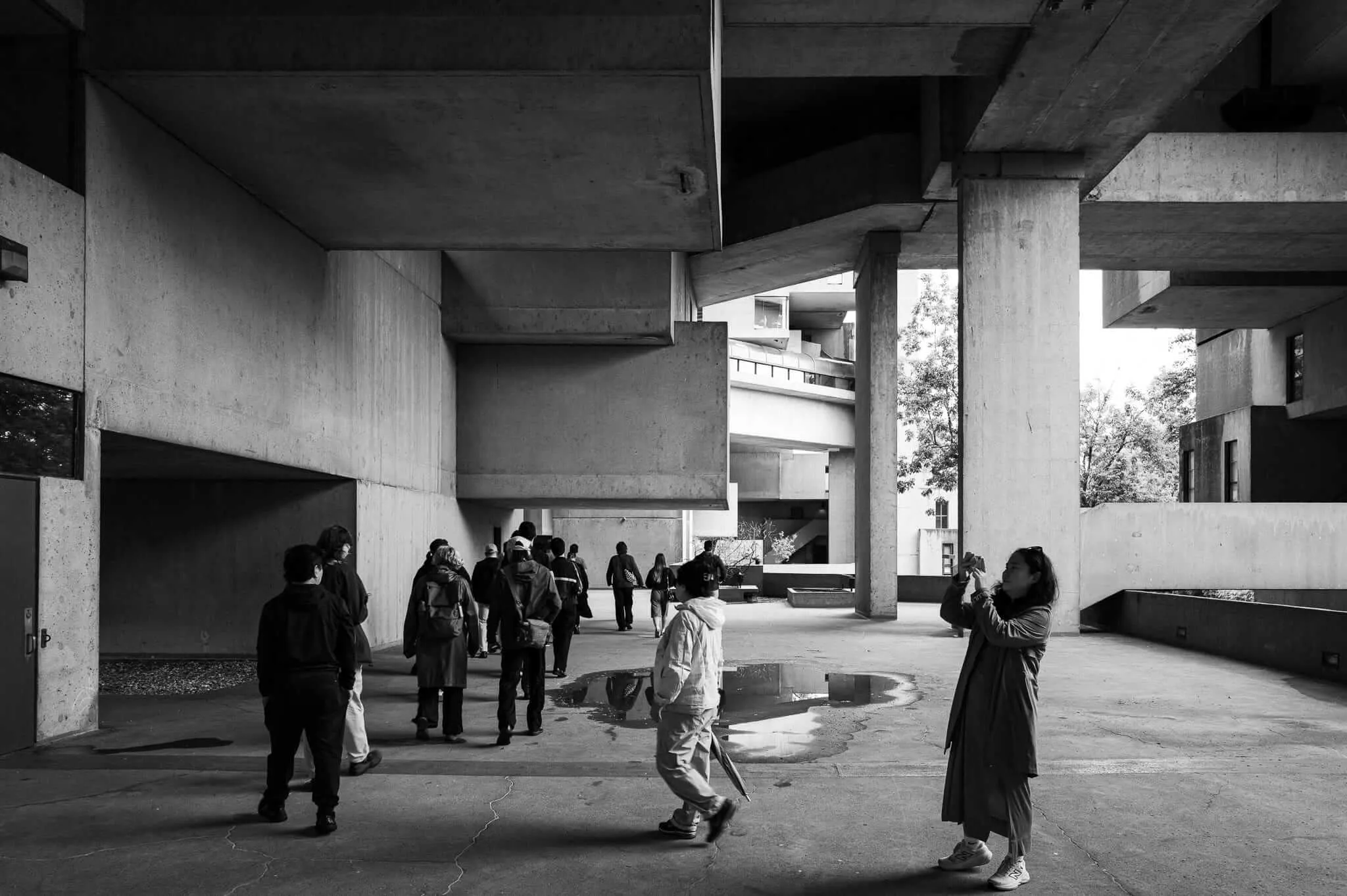 A group of visitors explore the dramatic piloti-supported undercroft of a brutalist concrete building, while a woman photographs the structure with her phone. Puddles reflect the massive columns in this black and white image.
