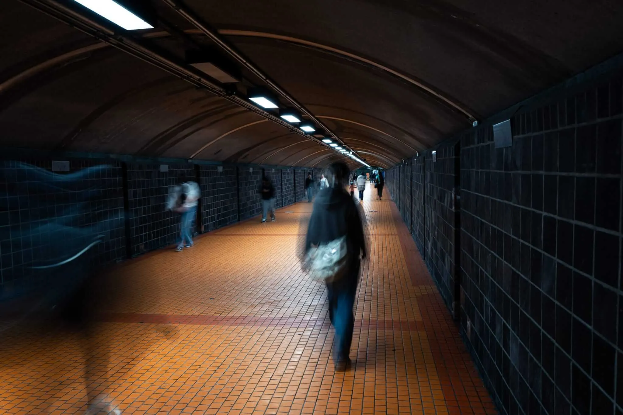 A long exposure photograph taken inside a pedestrian tunnel with an arched brick ceiling and warm orange tiled floor. Motion-blurred figures walk through the dimly lit underground passage, illuminated by a row of fluorescent ceiling lights receding into the distance. The closest figure carries a bag and appears ghost-like due to the long shutter speed.