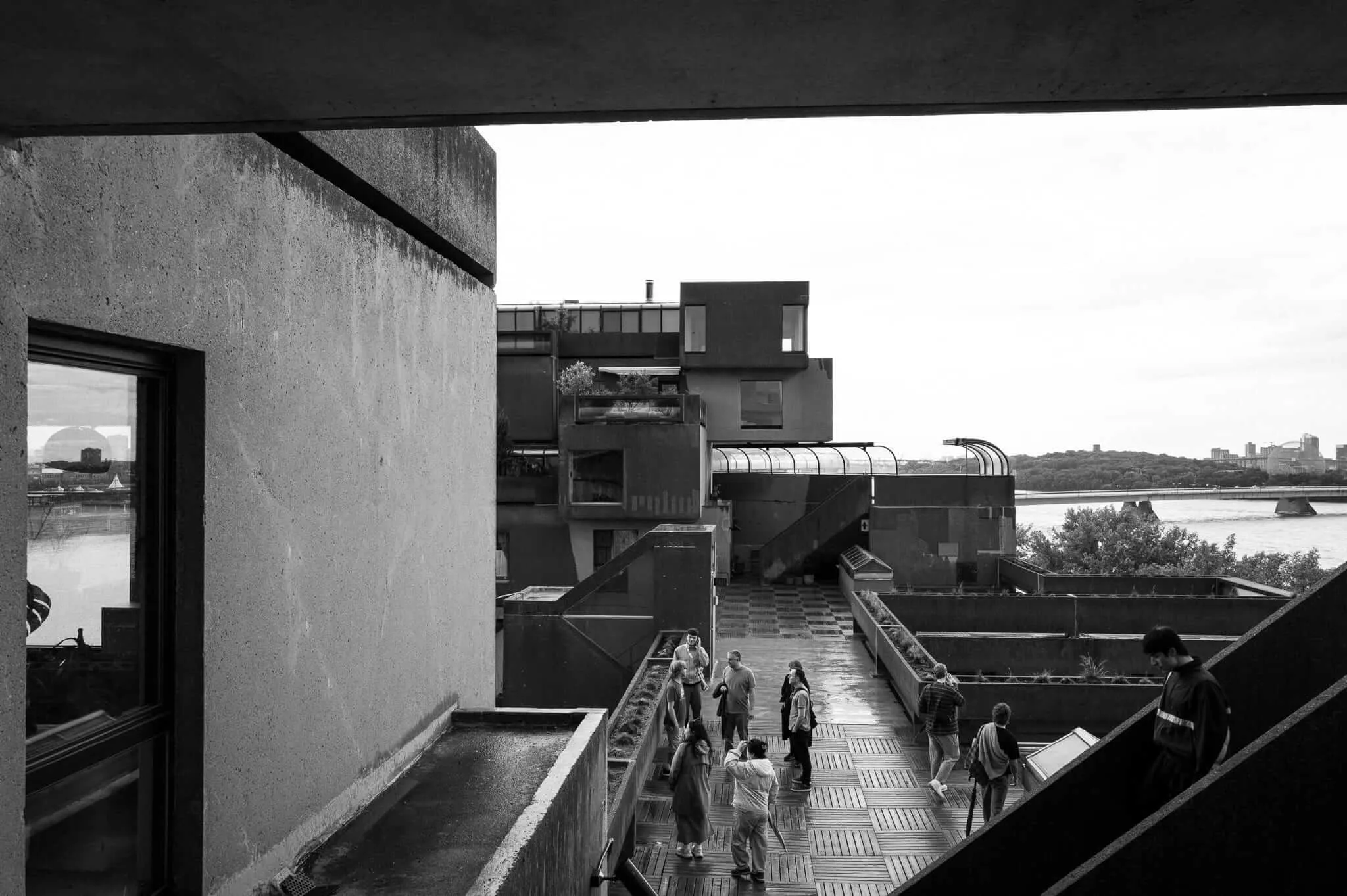 Black and white photograph taken from inside Habitat 67 in Montreal, looking out across a terraced concrete walkway where small groups of visitors wander and converse. The stacked modular brutalist housing units rise in the background, with the St. Lawrence River and distant city skyline visible beyond. A concrete overhang frames the top of the composition.