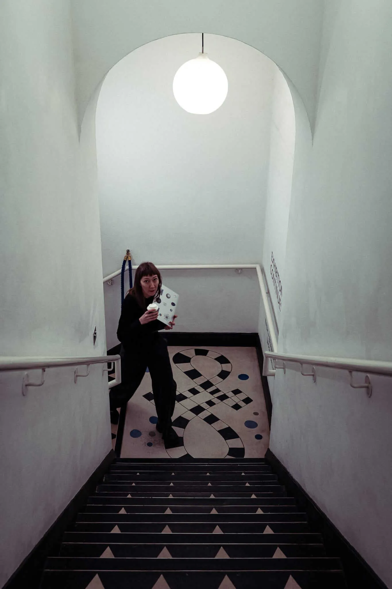 A woman with brown hair walking up the stairs, holding her laptop. She's positioned above black stairs with white triangular patterns, while below her is a large black and white checkered pattern on the floor. The space has white walls, handrails, and is lit by a single spherical pendant light hanging from an arched ceiling.