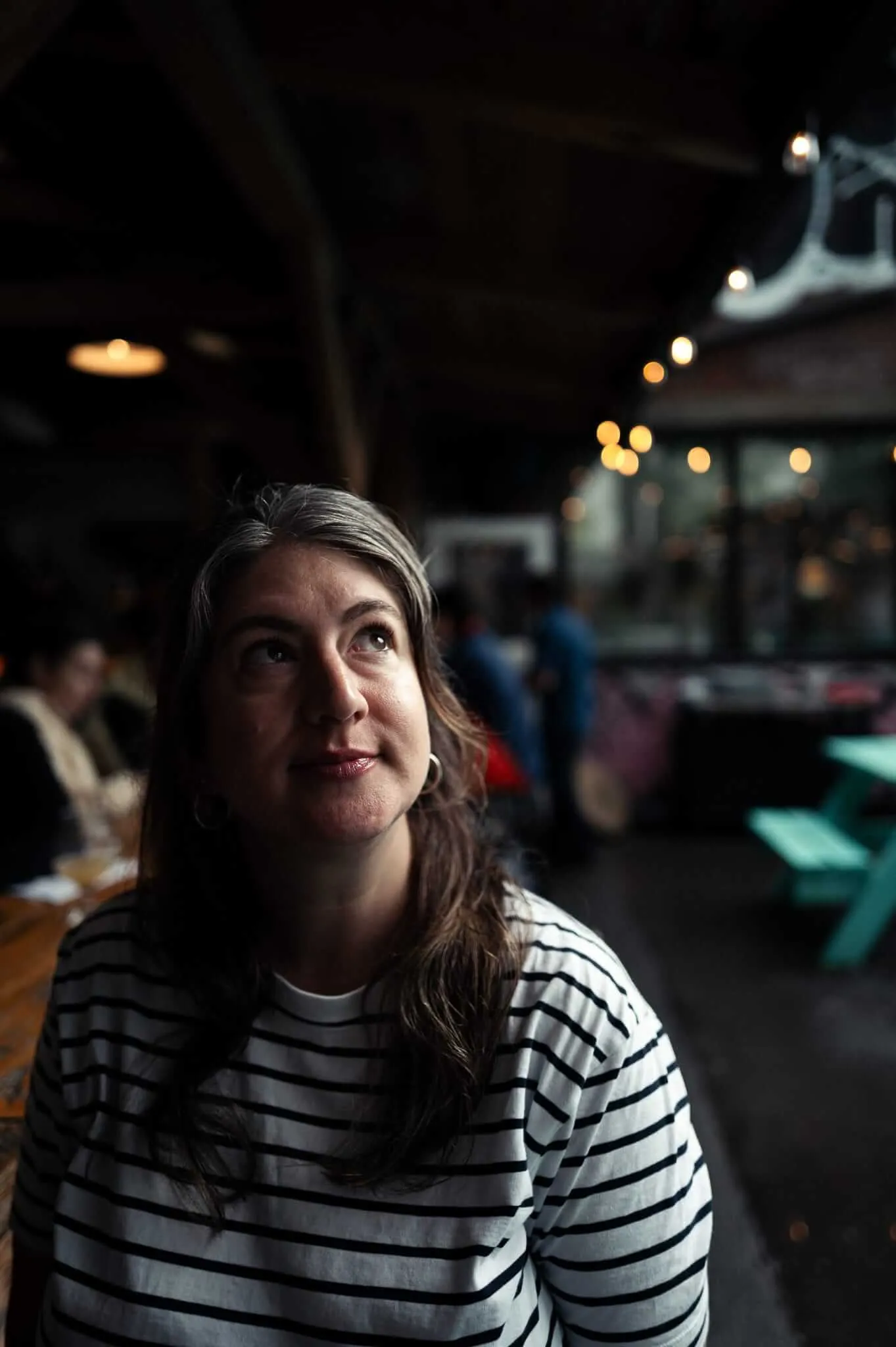 A woman with dark hair streaked with grey, wearing a white and black striped long-sleeve shirt, sits at a wooden table in a dimly lit outdoor venue. She gazes upward with a contemplative expression, illuminated by dramatic side lighting. Warm string lights and blurred figures populate the background.