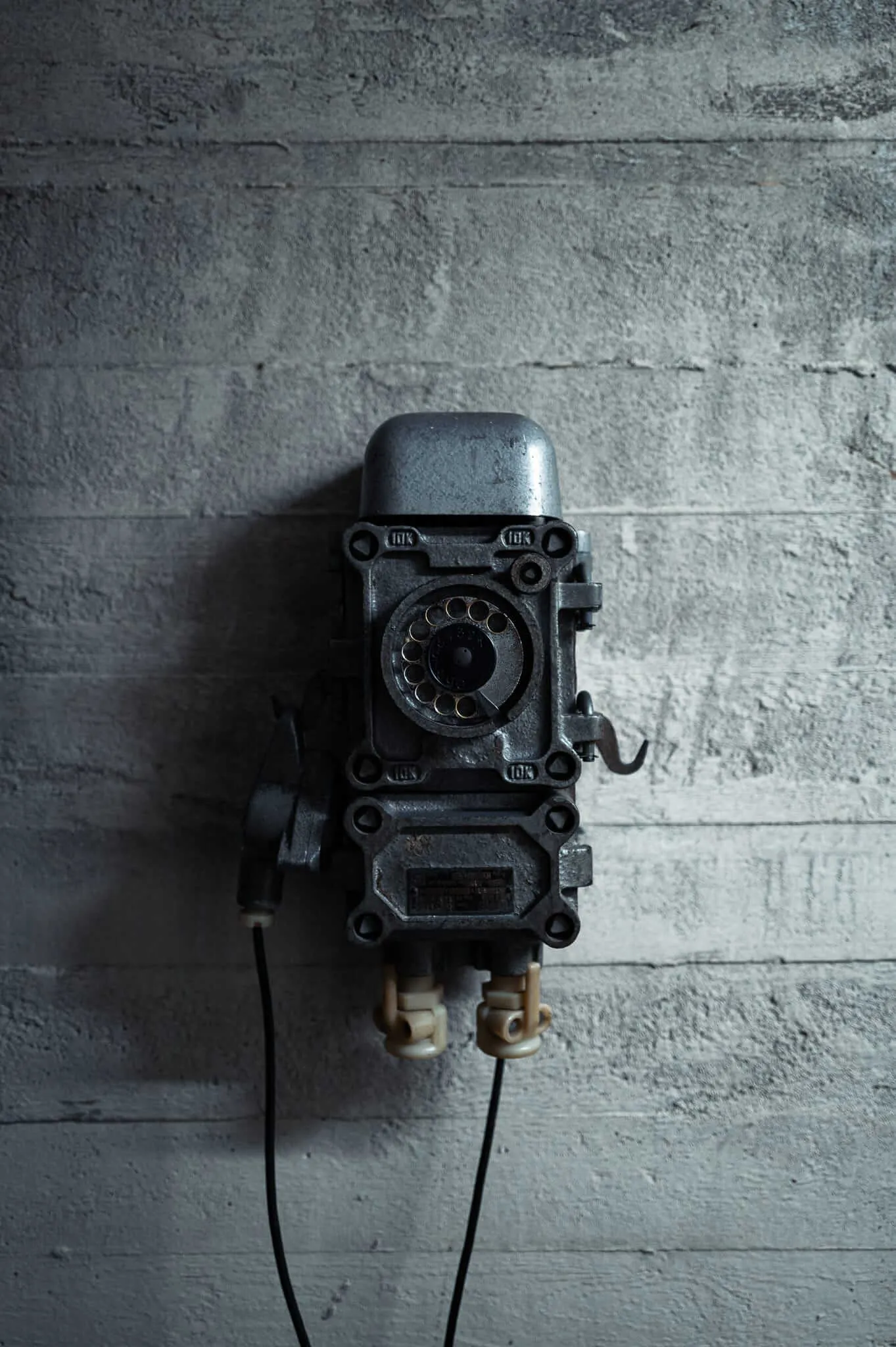 A vintage black rotary telephone mounted on a weathered concrete wall, with cream-colored receiver handset hanging below and dark cords extending downward