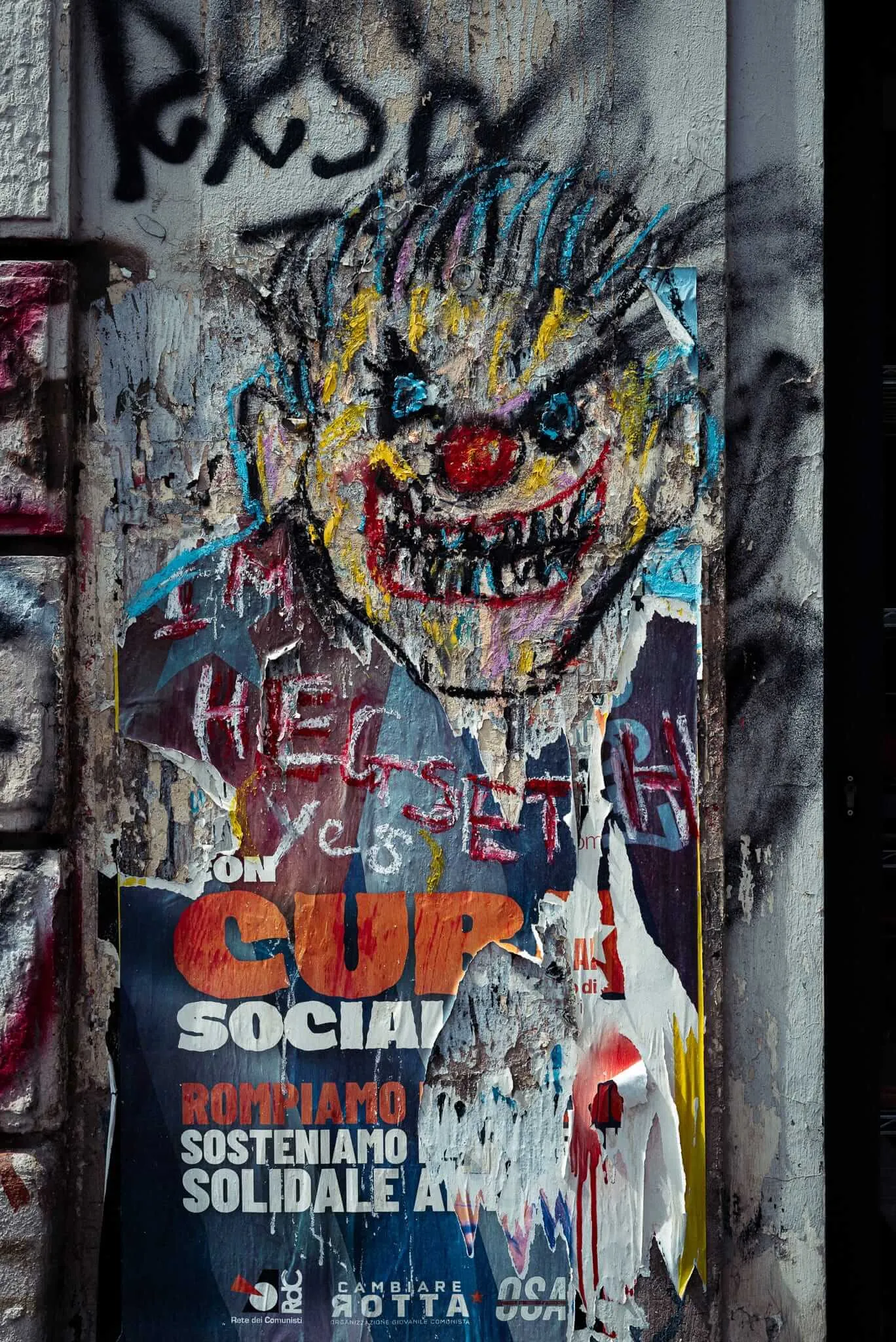 A weathered concrete wall covered in layered graffiti and street art, featuring a demonic cartoon face with wild hair and sharp teeth painted in bright colors, overlaid on torn political posters including one that reads 'CUP SOCIAL' in Italian. The artwork shows multiple layers of urban expression, with various tags, drawings, and text in different languages creating a palimpsest of street culture.