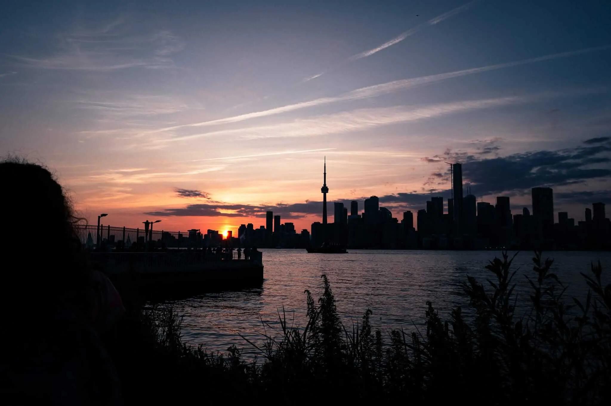 Silhouette of the Toronto skyline including the CN Tower viewed across Lake Ontario at sunset, with a figure in the foreground and dramatic orange and blue sky with wispy clouds overhead.