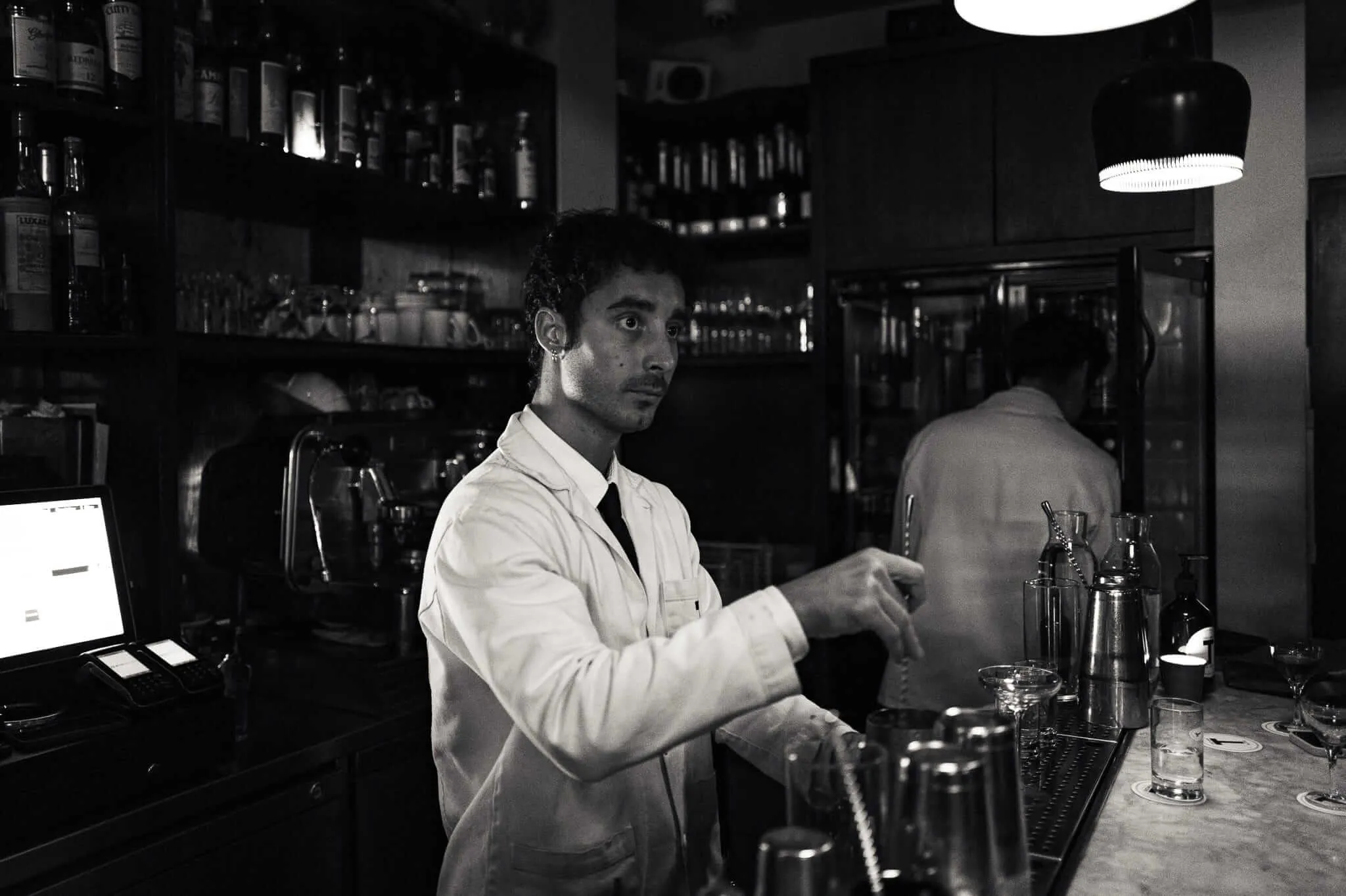 Black and white photograph of a bartender in white shirt and tie working behind a bar, with shelves of bottles visible in the background and pendant lighting overhead.