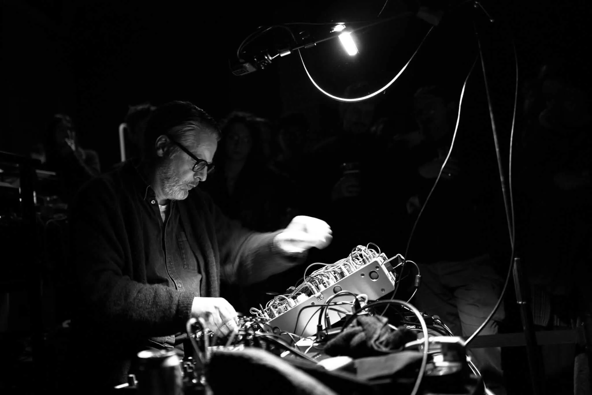 Black and white photograph of a bearded man with glasses wearing dark clothing, hunched over an electronic music setup with synthesizers, cables, and equipment on a table under dramatic lighting in what appears to be a performance or studio space