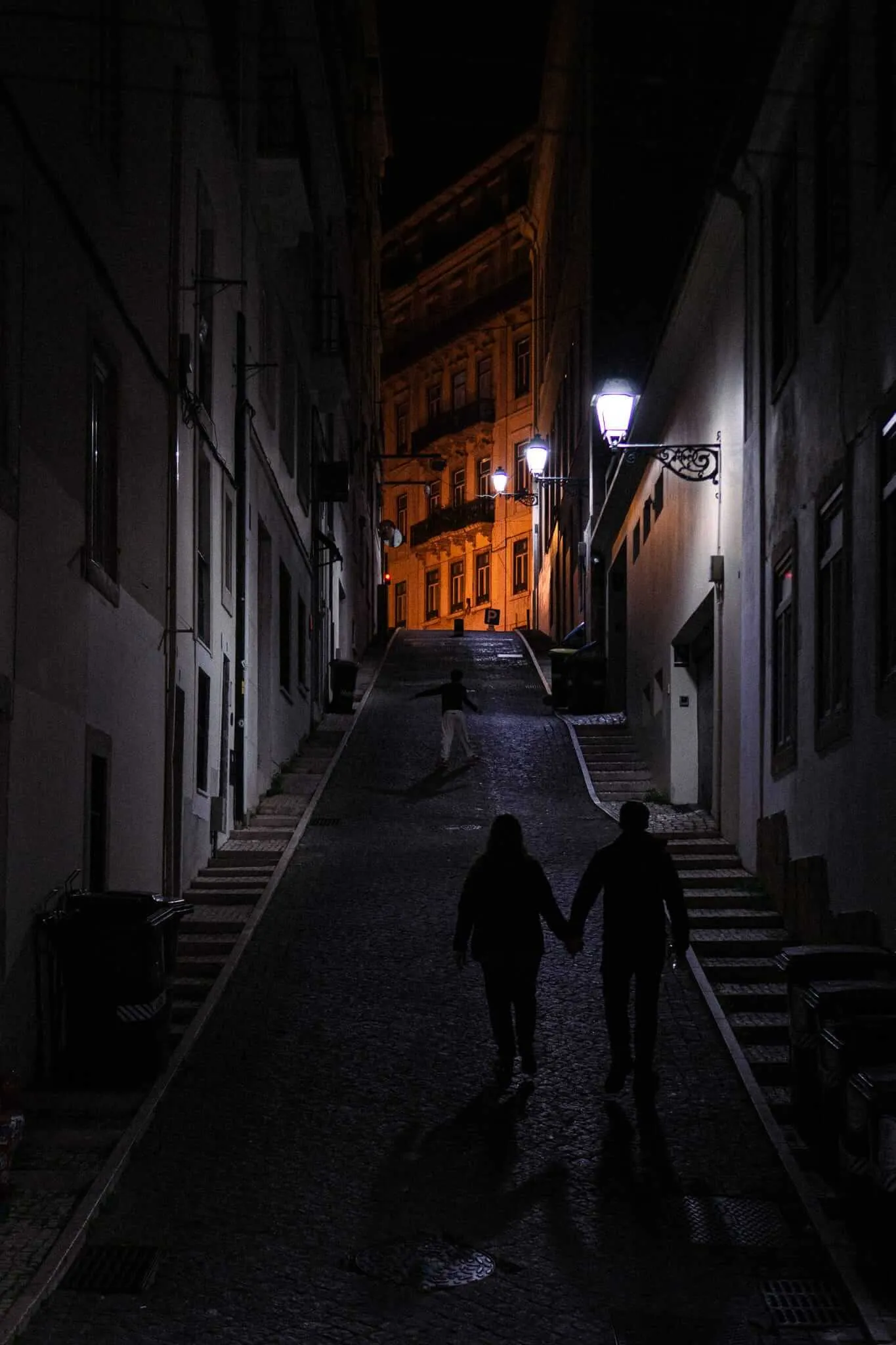 Two silhouetted figures hold hands while walking up a narrow cobblestone alley at night, illuminated by ornate street lamps, with a warmly lit historic building glowing orange in the distance.