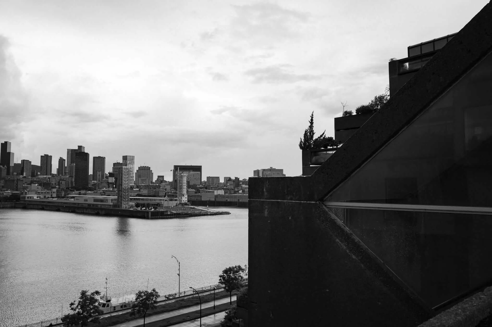 Black and white photograph taken from an elevated position beside a striking brutalist concrete building with angular stepped terraces and rooftop vegetation, looking out across a wide river toward a dense urban skyline under a cloudy sky. A small boat and waterfront promenade are visible in the foreground below.