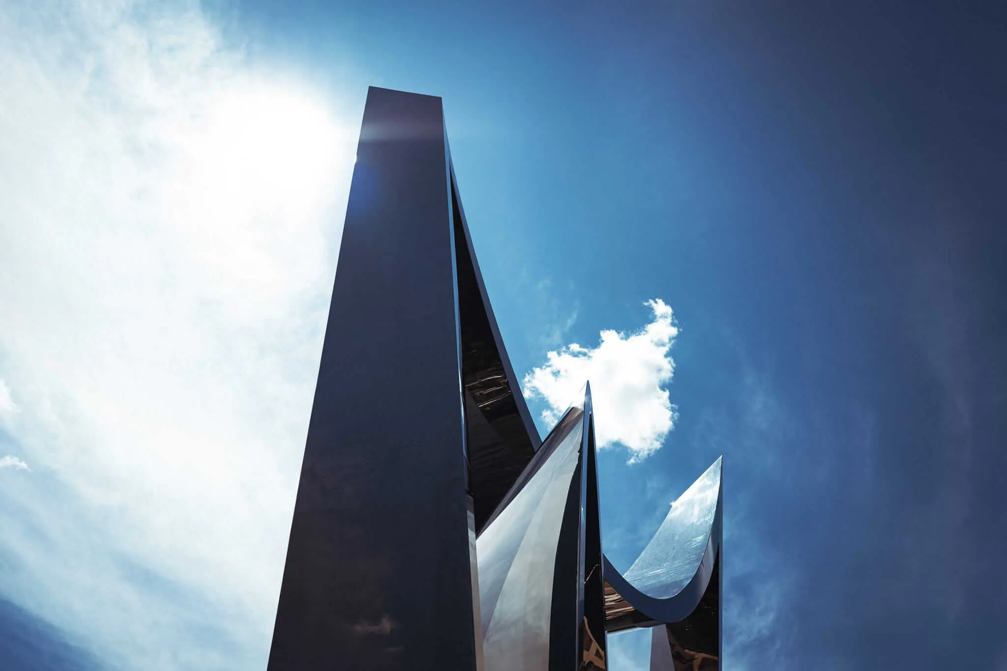 A low-angle view looking up at a towering abstract metal sculpture with sharp, blade-like forms and polished reflective surfaces, set against a vivid blue sky with scattered clouds and bright sunlight.