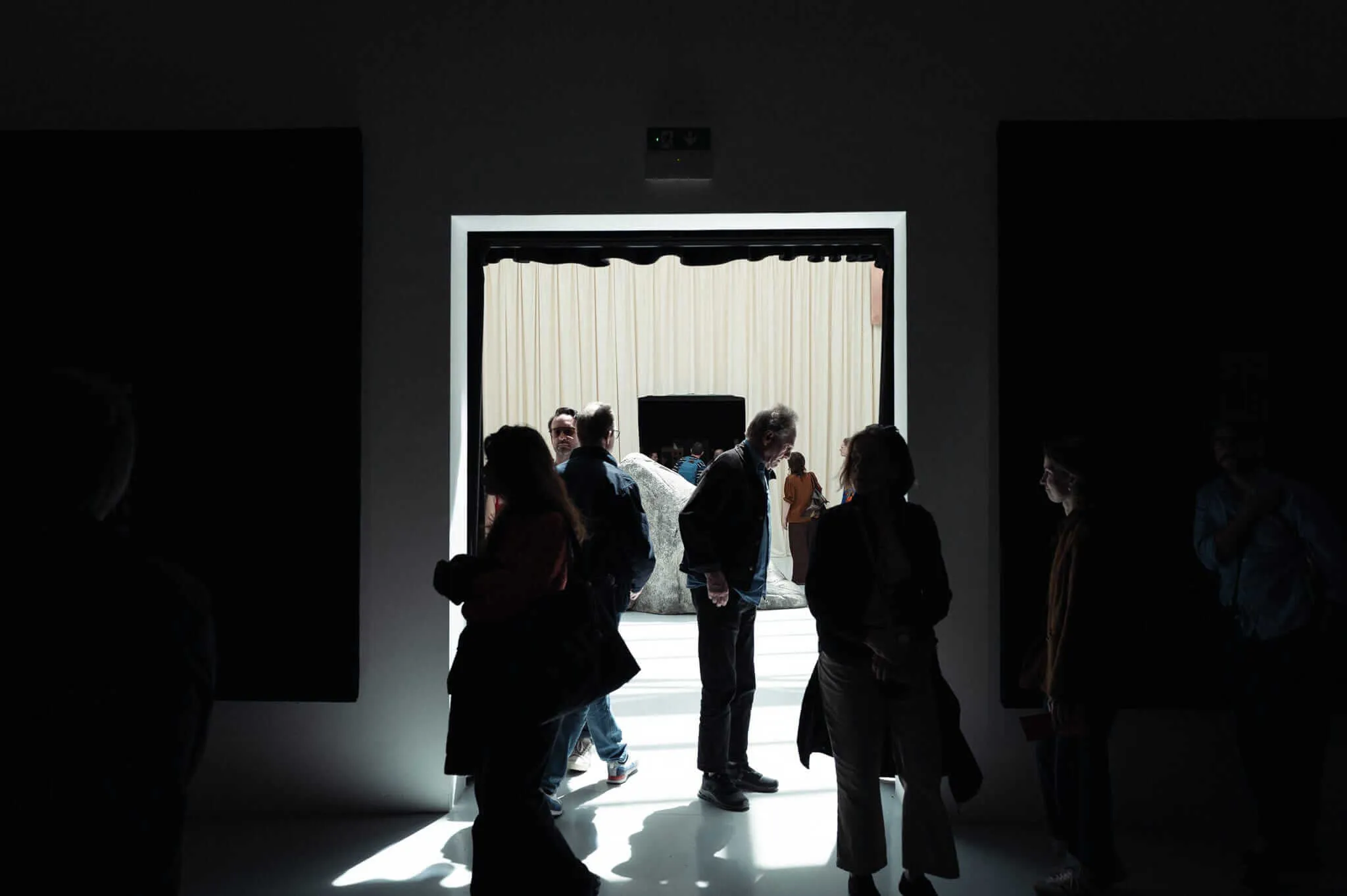Visitors in silhouette stand in a darkened gallery space before a brightly lit doorway, beyond which a large sculptural form and cream curtains are visible in an adjacent exhibition room.