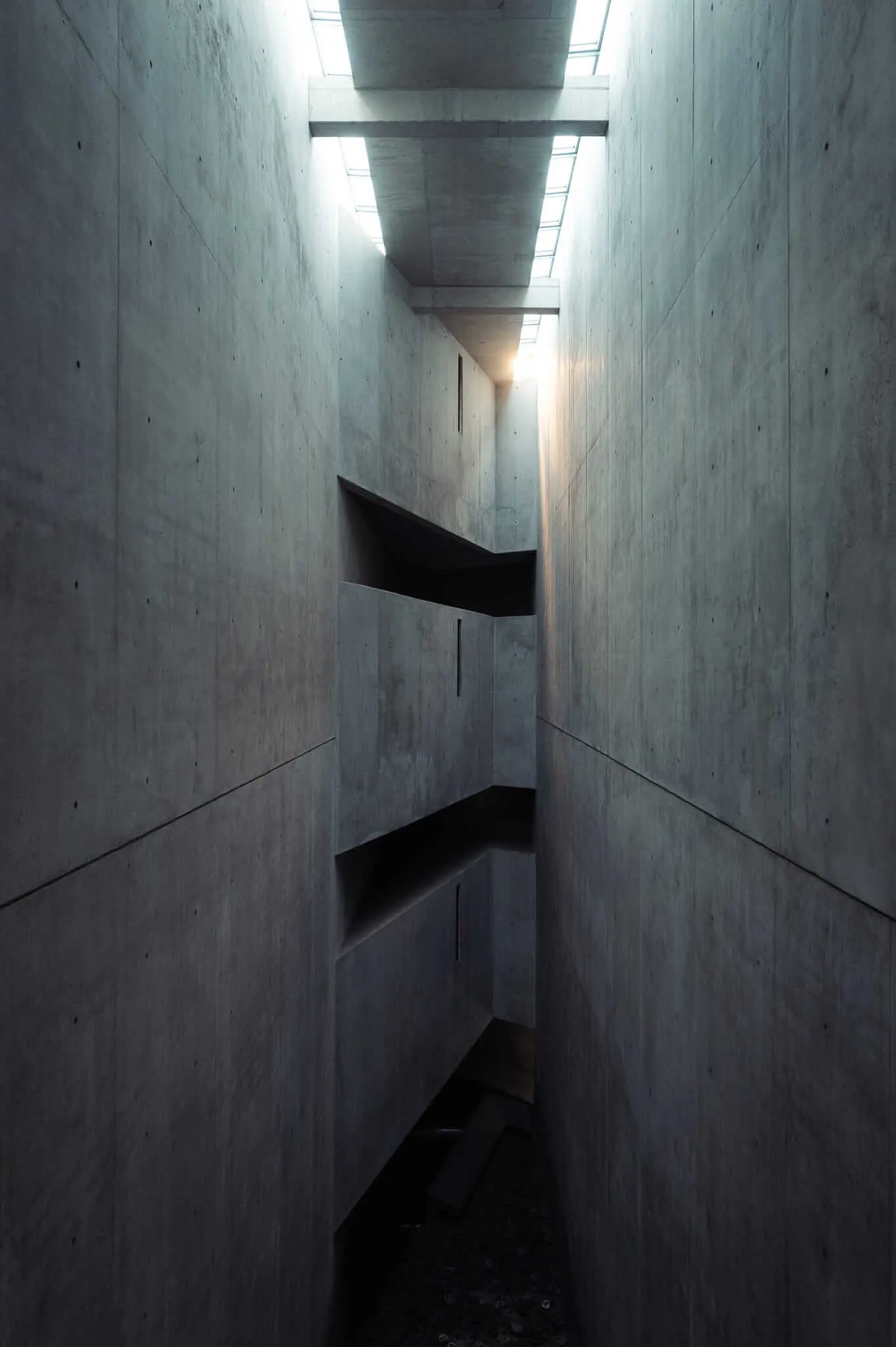 A stark concrete interior featuring multiple levels connected by dark geometric platforms or bridges, with natural light filtering down from skylights above, creating dramatic shadows and highlighting the raw concrete textures and brutalist architectural forms