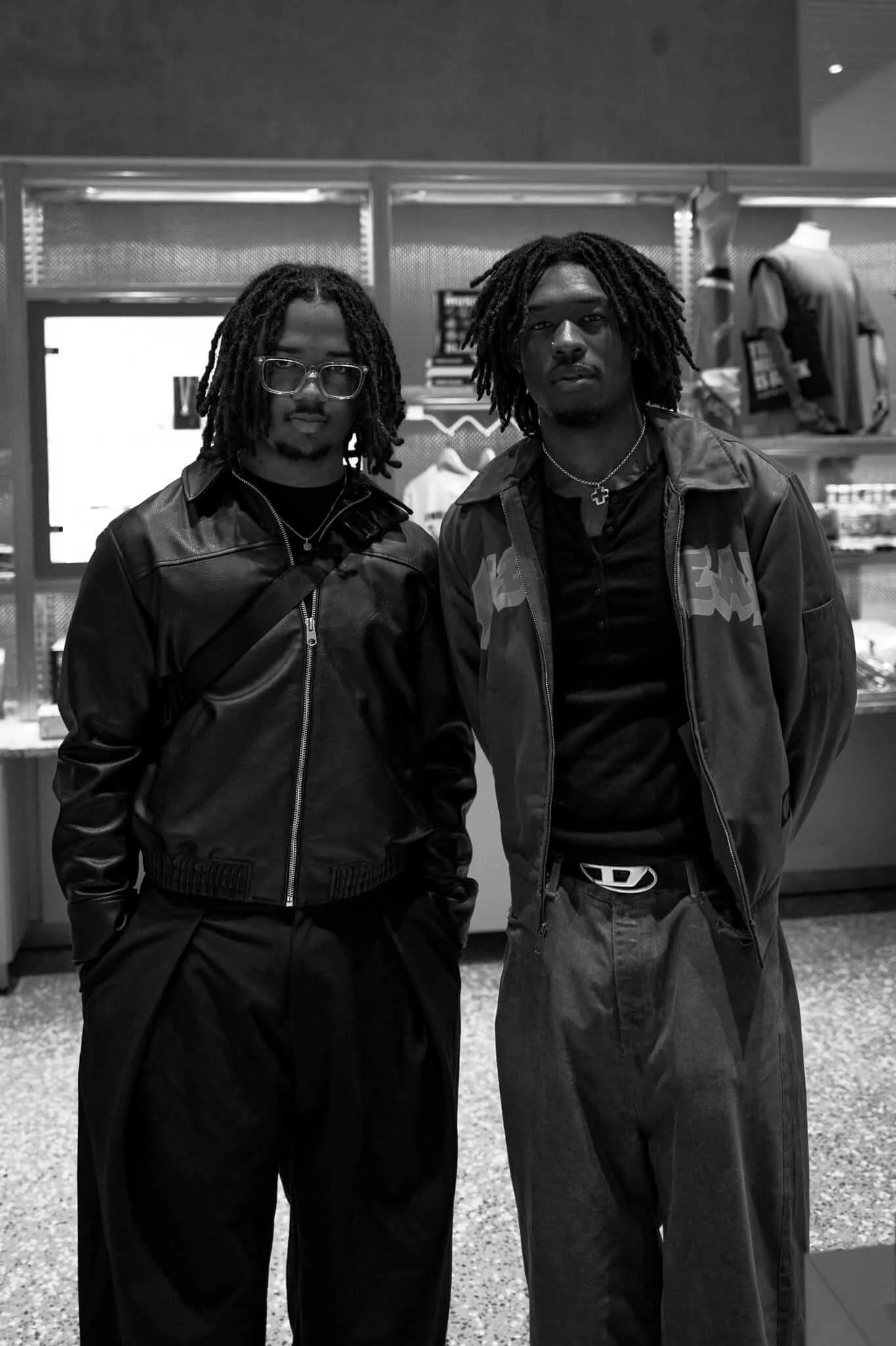 Black and white photograph of two young men with dreadlocks standing side by side in a museum gift shop. The man on the left wears glasses and a leather jacket, while the man on the right wears a longer coat with text on it. Behind them are illuminated storefronts and display cases. The composition creates a sense of solidarity and shared presence in an urban commercial environment.