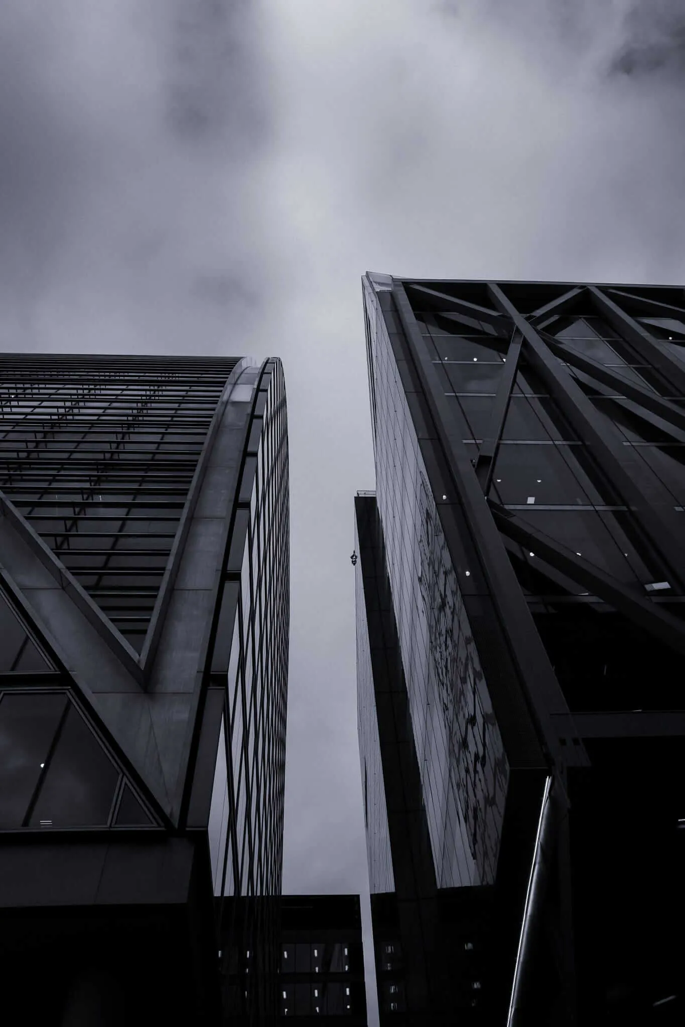 Dramatic low-angle view of modern skyscrapers with glass and steel facades featuring geometric patterns and exposed structural elements, shot against a moody cloudy sky in monochromatic tones