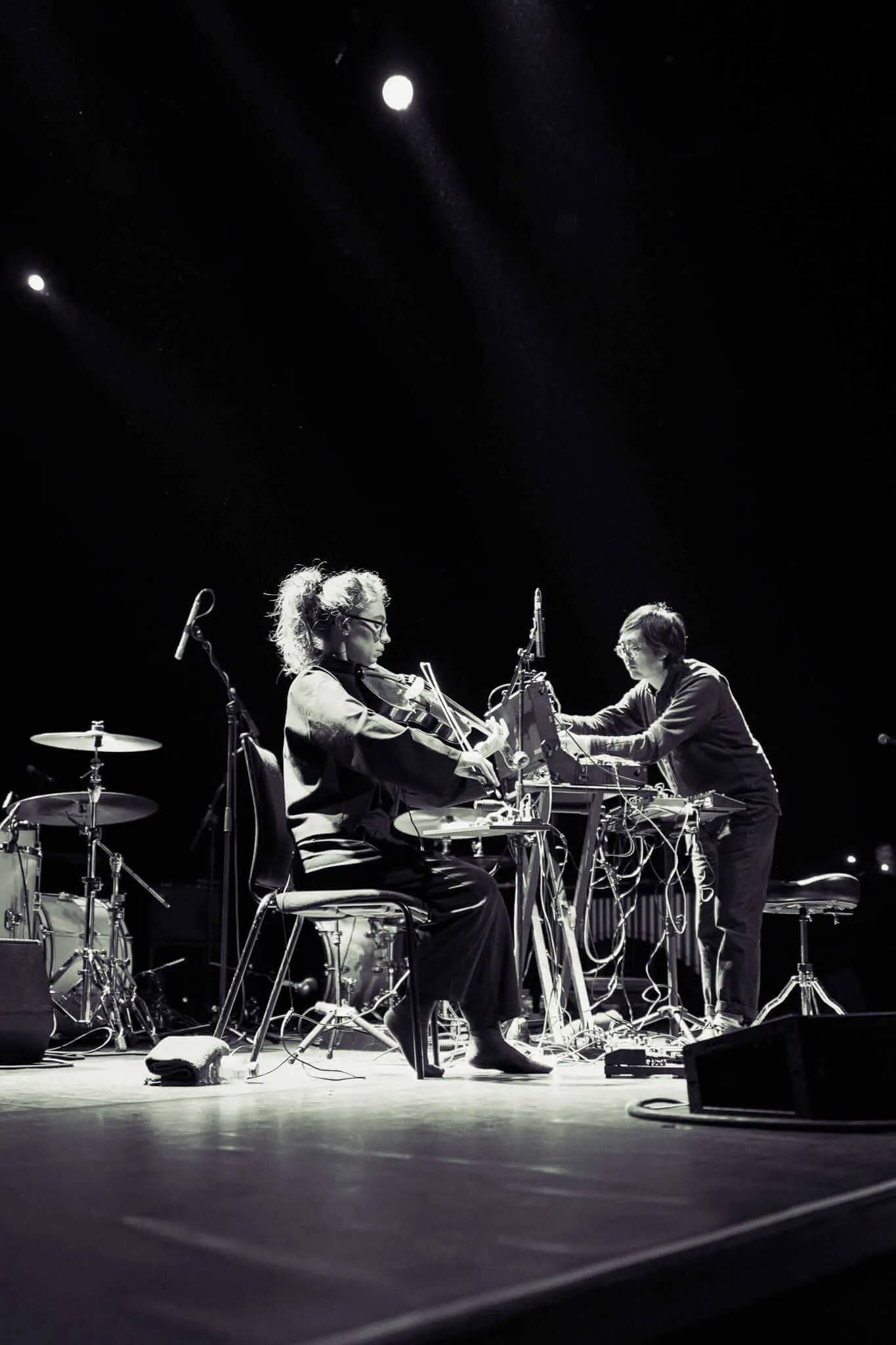 Black and white photograph of two musicians performing on a dimly lit stage, with one seated playing violin while another stands at electronic equipment, surrounded by drum kit and various musical instruments under dramatic spotlighting