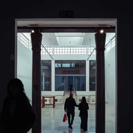 A silhouetted figure in the foreground watches as two visitors walk through a grand doorway flanked by granite columns into a brightly lit museum atrium displaying an Ettore Sottsass exhibition, with stairs and glass walls visible in the background.