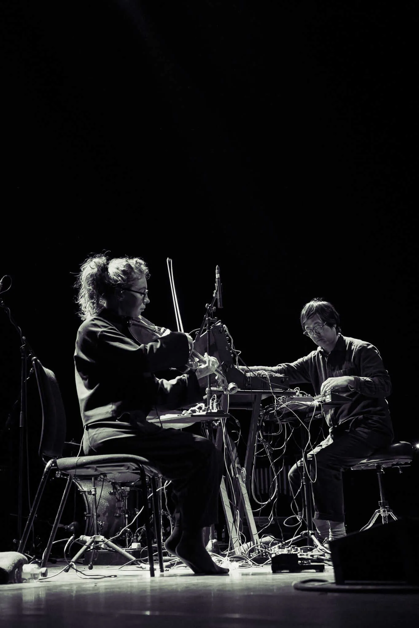 Black and white photograph of two musicians performing on stage - one playing violin on the left, another working with electronic equipment on the right, surrounded by cables, microphones, and musical gear against a dark background