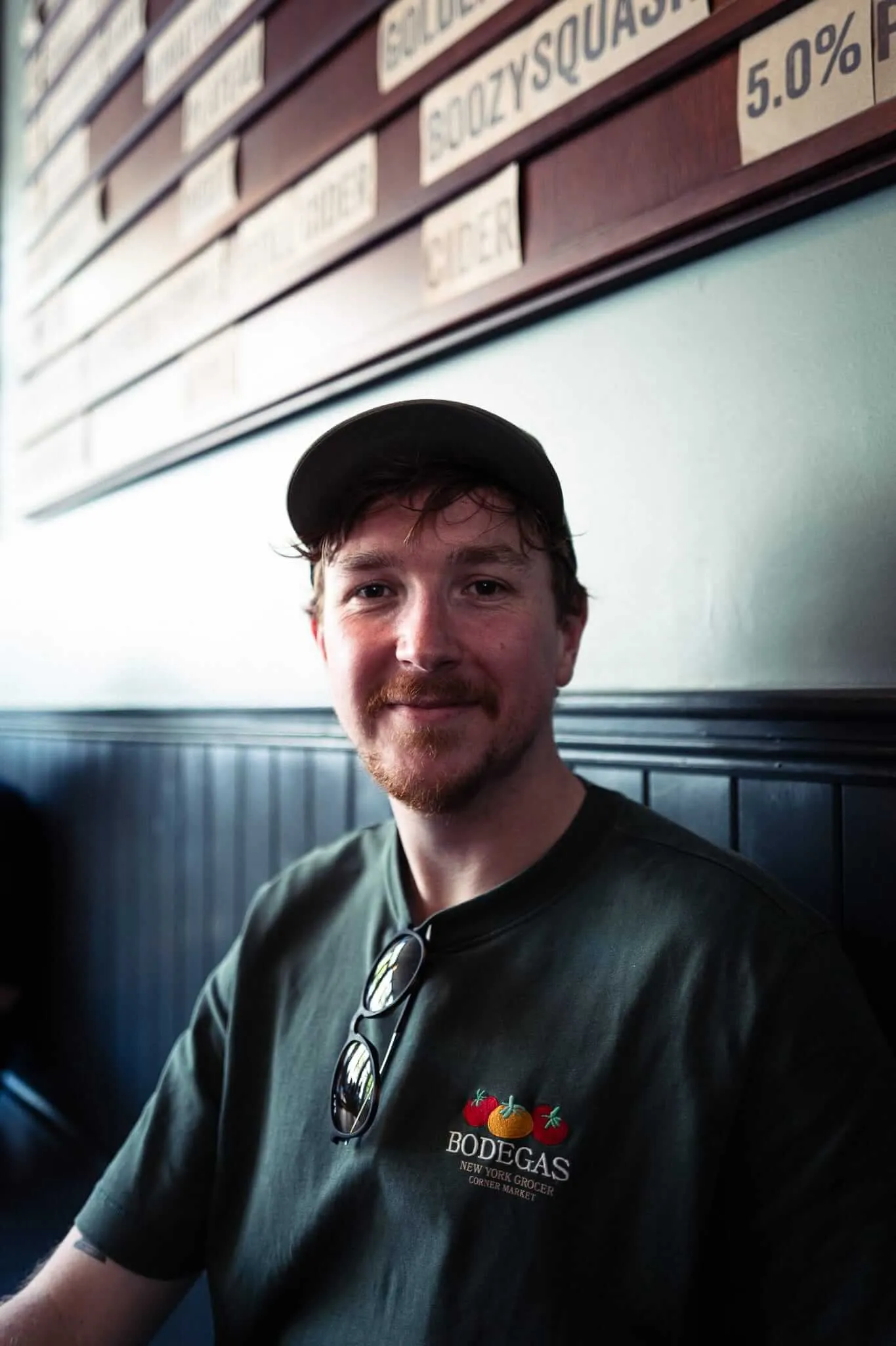 A bearded man wearing a black baseball cap and green Bodegas t-shirt stands in what appears to be a grocery store or market, with product shelves visible behind him. He has a slight smile and is looking directly at the camera.