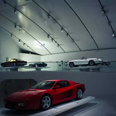 A red Ferrari sports car displayed on a white platform in the foreground of a sleek museum gallery, with several classic cars arranged on an elevated mezzanine level behind a glass partition, all illuminated by dramatic track lighting against curved white walls.