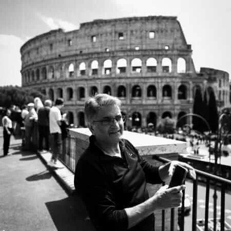 A middle-aged man wearing glasses and a black shirt holds a smartphone while standing in front of the Colosseum in Rome, surrounded by other tourists on a sunny day. The image is in black and white.