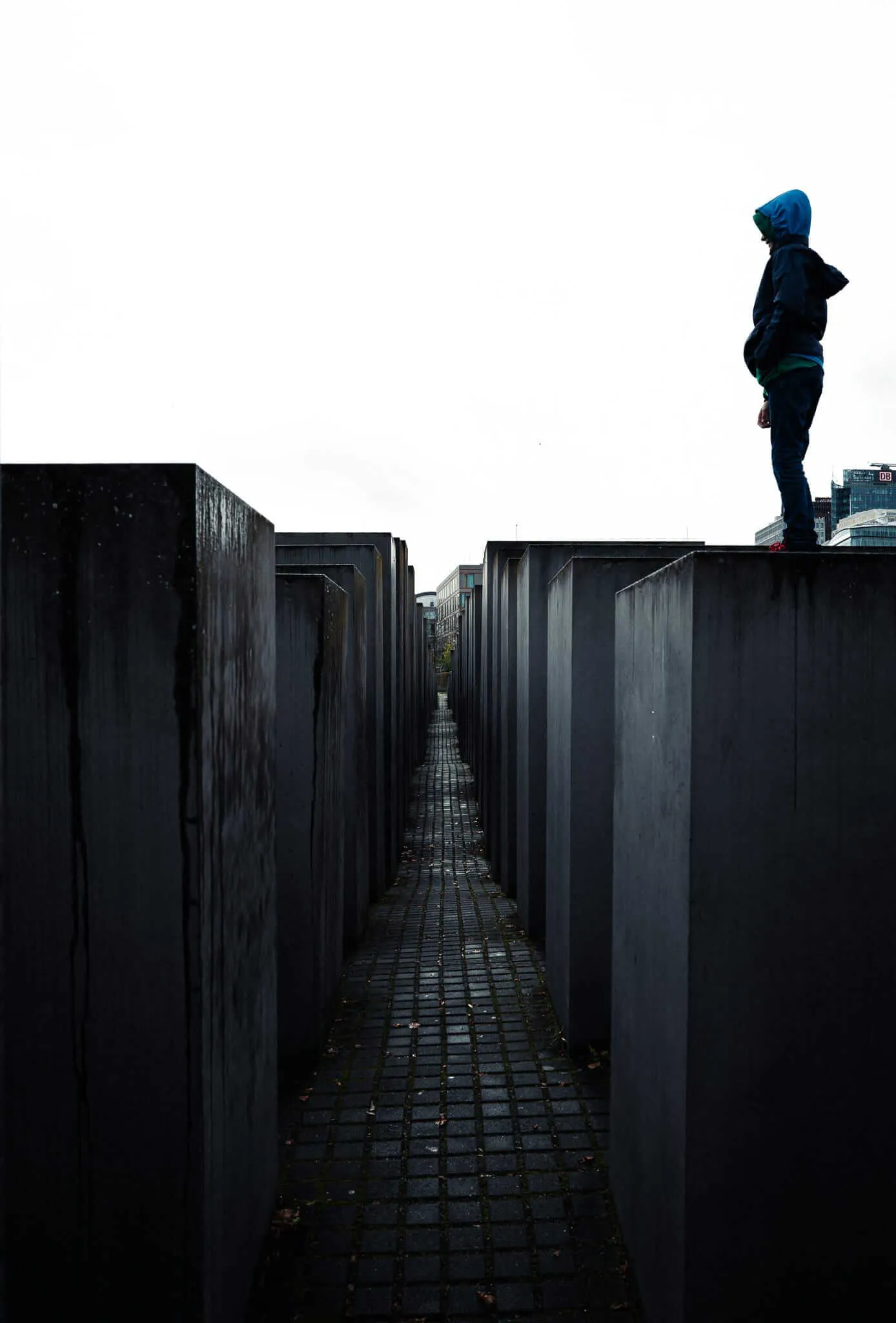 A lone figure in dark clothing and hood stands atop a tall concrete slab of the Holocaust Memorial in Berlin, silhouetted against an overcast sky. Below, narrow pathways wind between the gray concrete blocks of varying heights, creating a maze-like pattern that stretches toward the urban skyline in the distance.