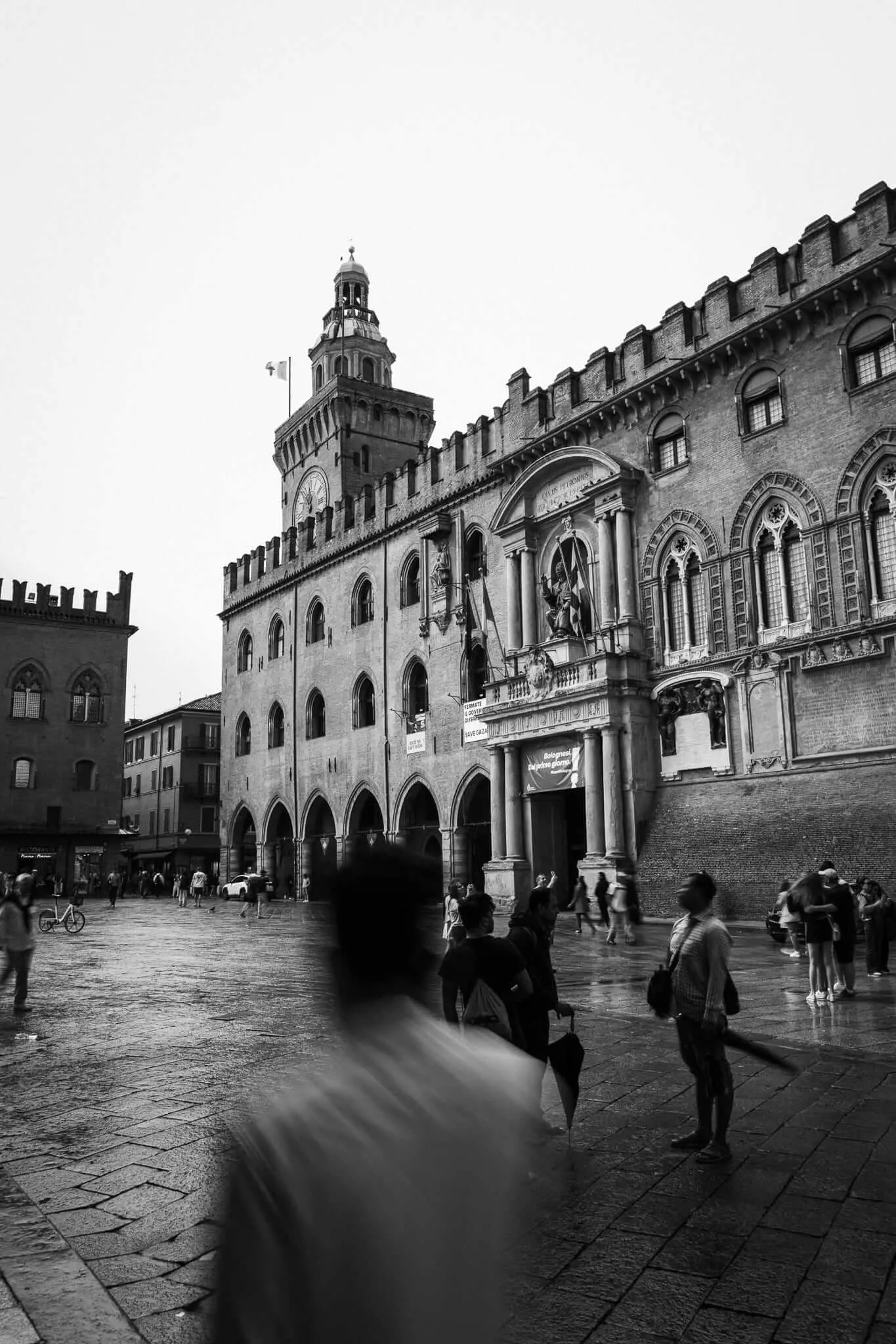 Black and white photograph of Bologna's Palazzo d'Accursio and Palazzo del Podestà in rainy weather, with a ghostly motion-blurred figure in the foreground and scattered pedestrians crossing the wet cobblestone piazza beneath a grey sky
