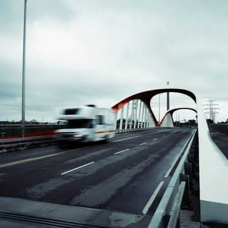 A motion-blurred white transport truck crosses a modern arch bridge with striking red and white steel arches under an overcast grey sky, with power transmission towers visible in the background.