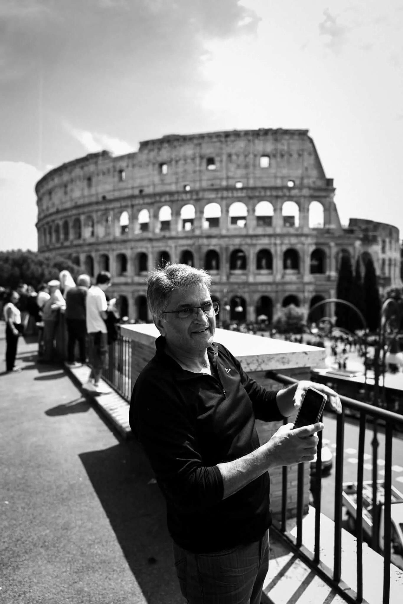 A middle-aged man wearing glasses and a black shirt holds a smartphone while standing in front of the Colosseum in Rome, surrounded by other tourists on a sunny day. The image is in black and white.