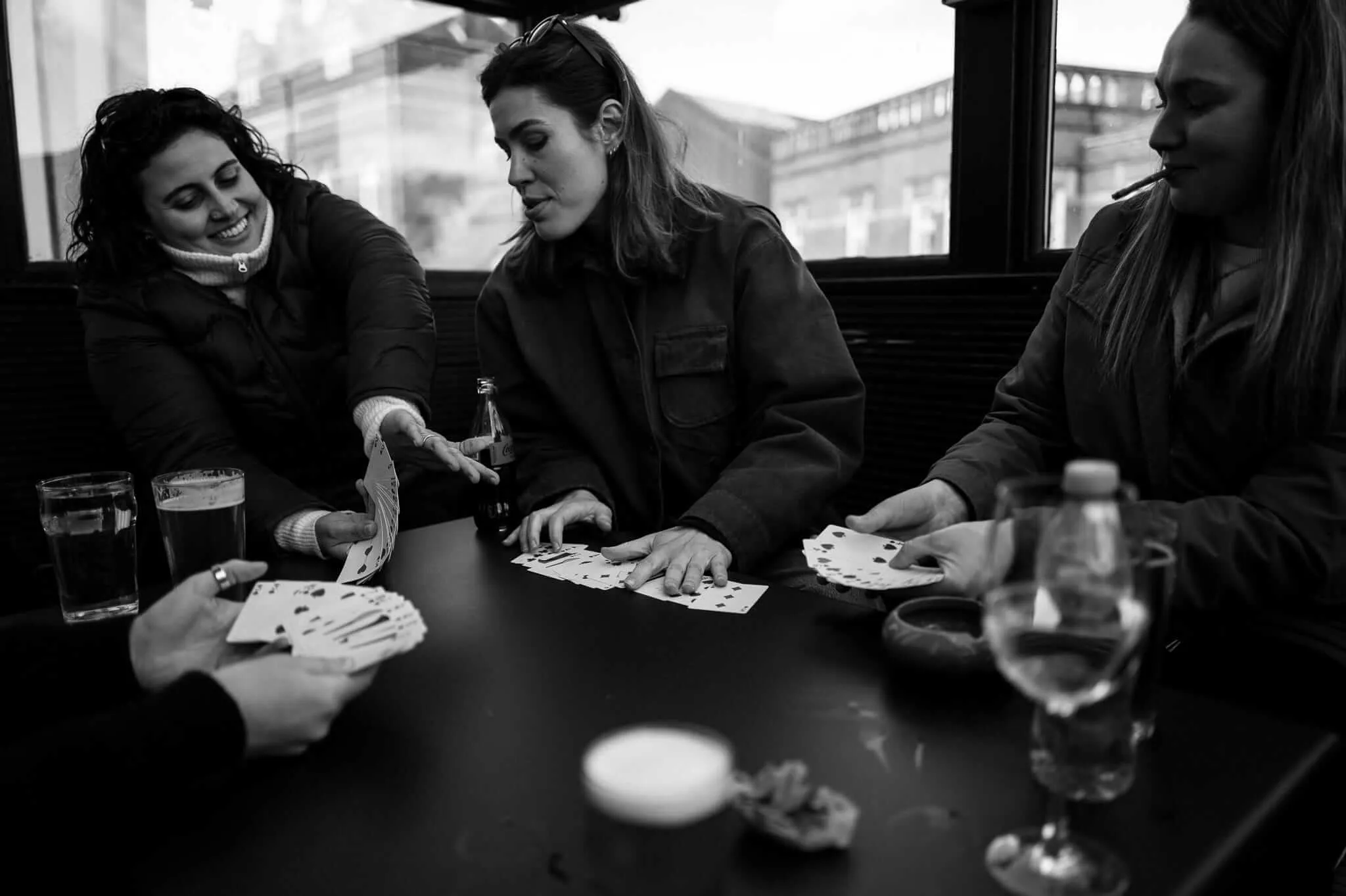 Black and white photograph of three women sitting at a table in what appears to be a pub or restaurant, playing cards together. The woman on the left is smiling and leaning back, while the middle woman deals cards. Various drinks including beer and wine glasses are visible on the dark wooden table. Large windows show urban buildings in the background.