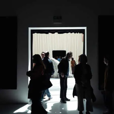 Visitors in silhouette stand in a darkened gallery space before a brightly lit doorway, beyond which a large sculptural form and cream curtains are visible in an adjacent exhibition room.